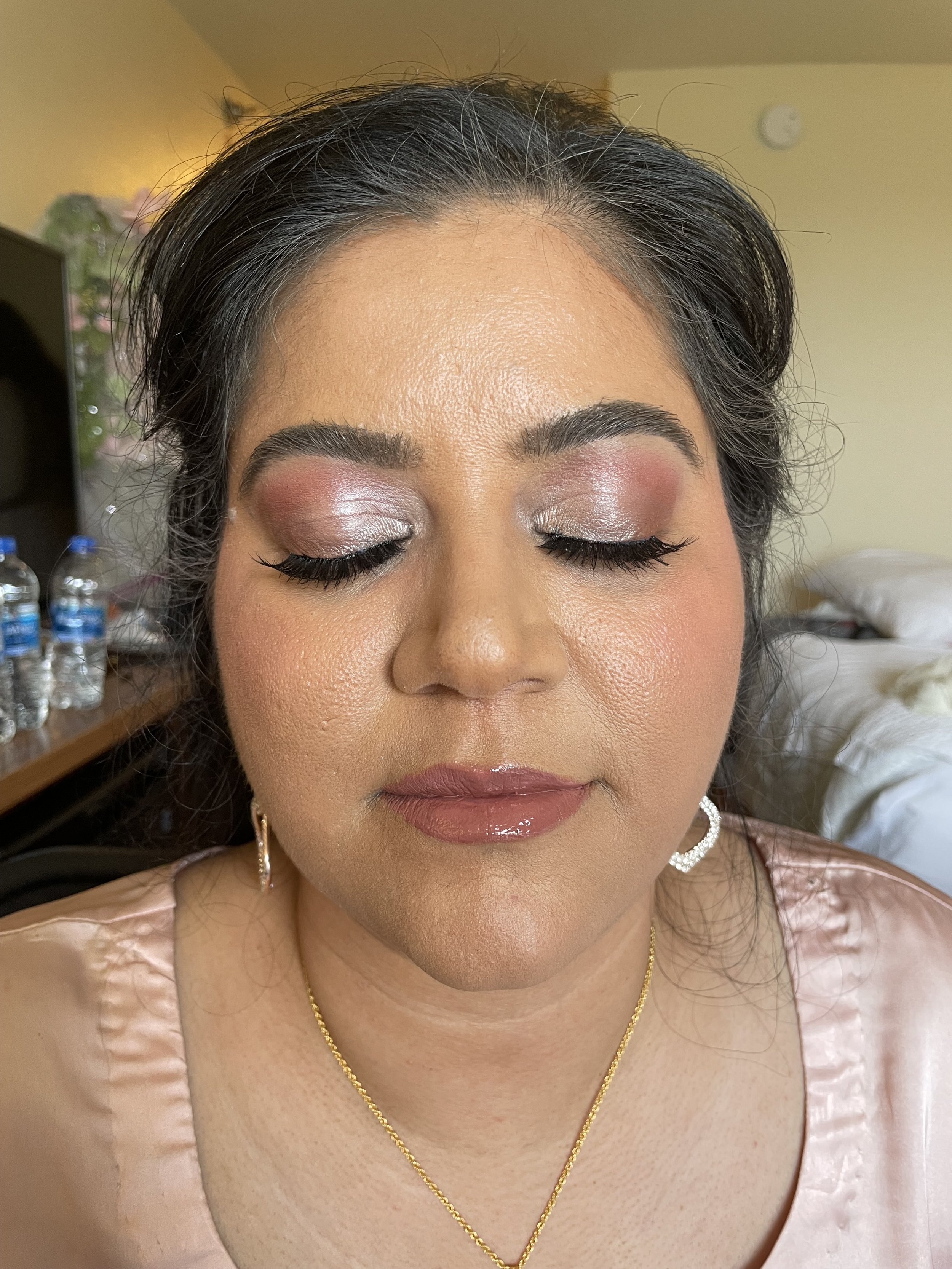 Close-up of a woman with closed eyes, wearing makeup, earrings, a gold necklace, and a light pink satin top, in a room.