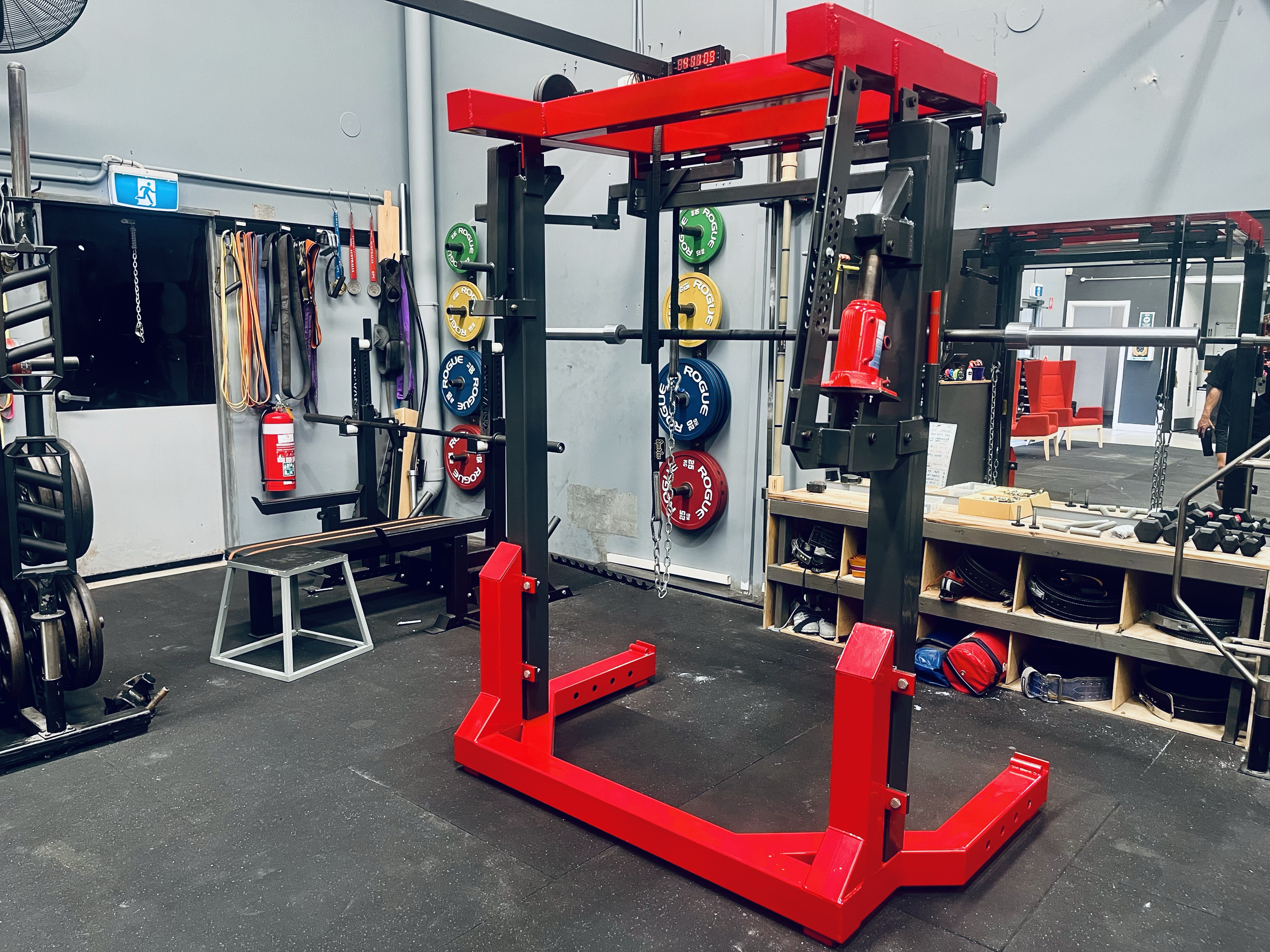 Power rack in a gym with weight plates on the wall and various workout equipment around.