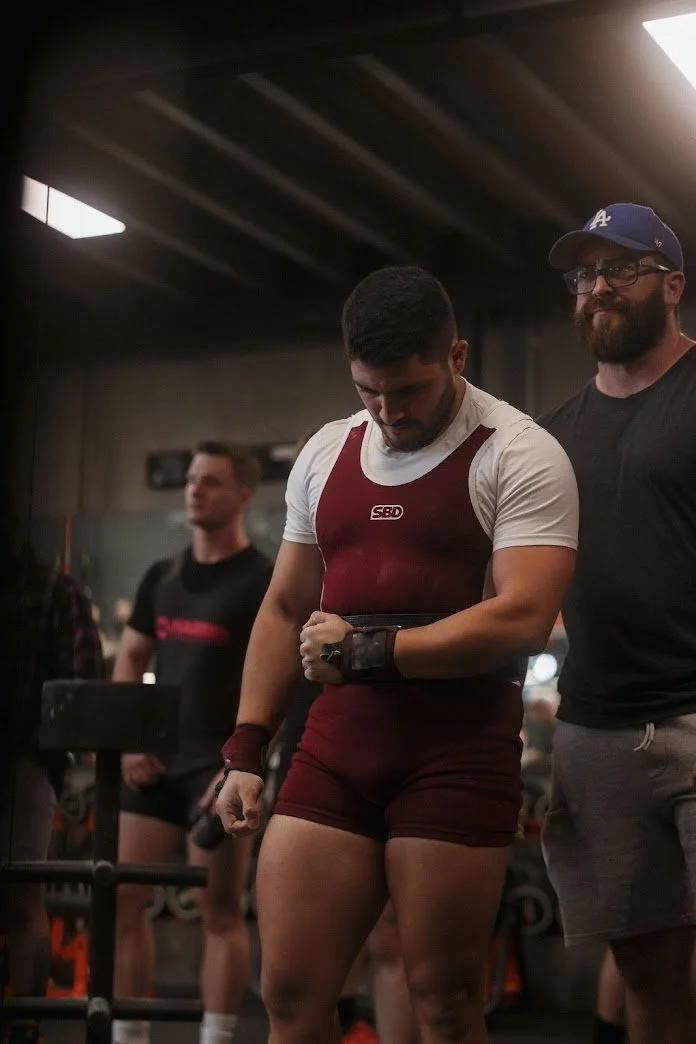 A weightlifter in a maroon singlet and white shirt preparing for a lift at the gym, standing between two men, one in a black shirt and blue cap, the other blurred in the background.