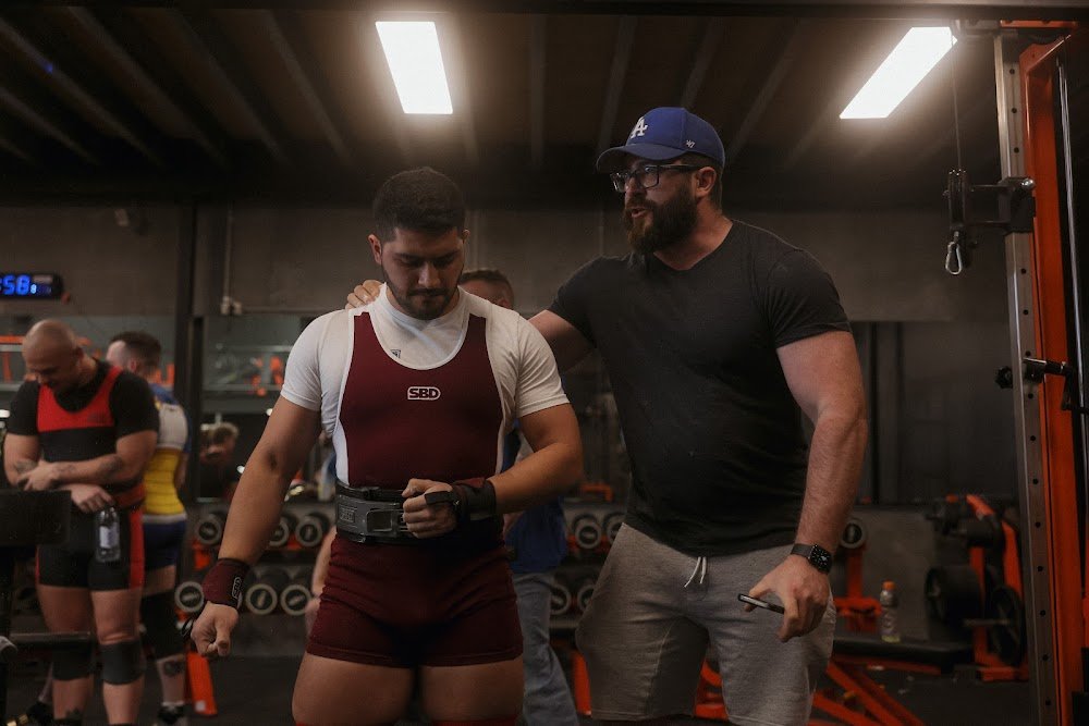 A coach giving instructions to a male powerlifter in a gym, surrounded by other athletes, with gym equipment and a wall clock in the background.