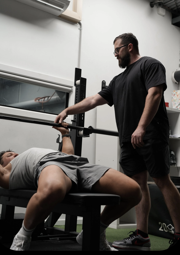 A man lying on a workout bench lifting weights while another man spots him in a gym.