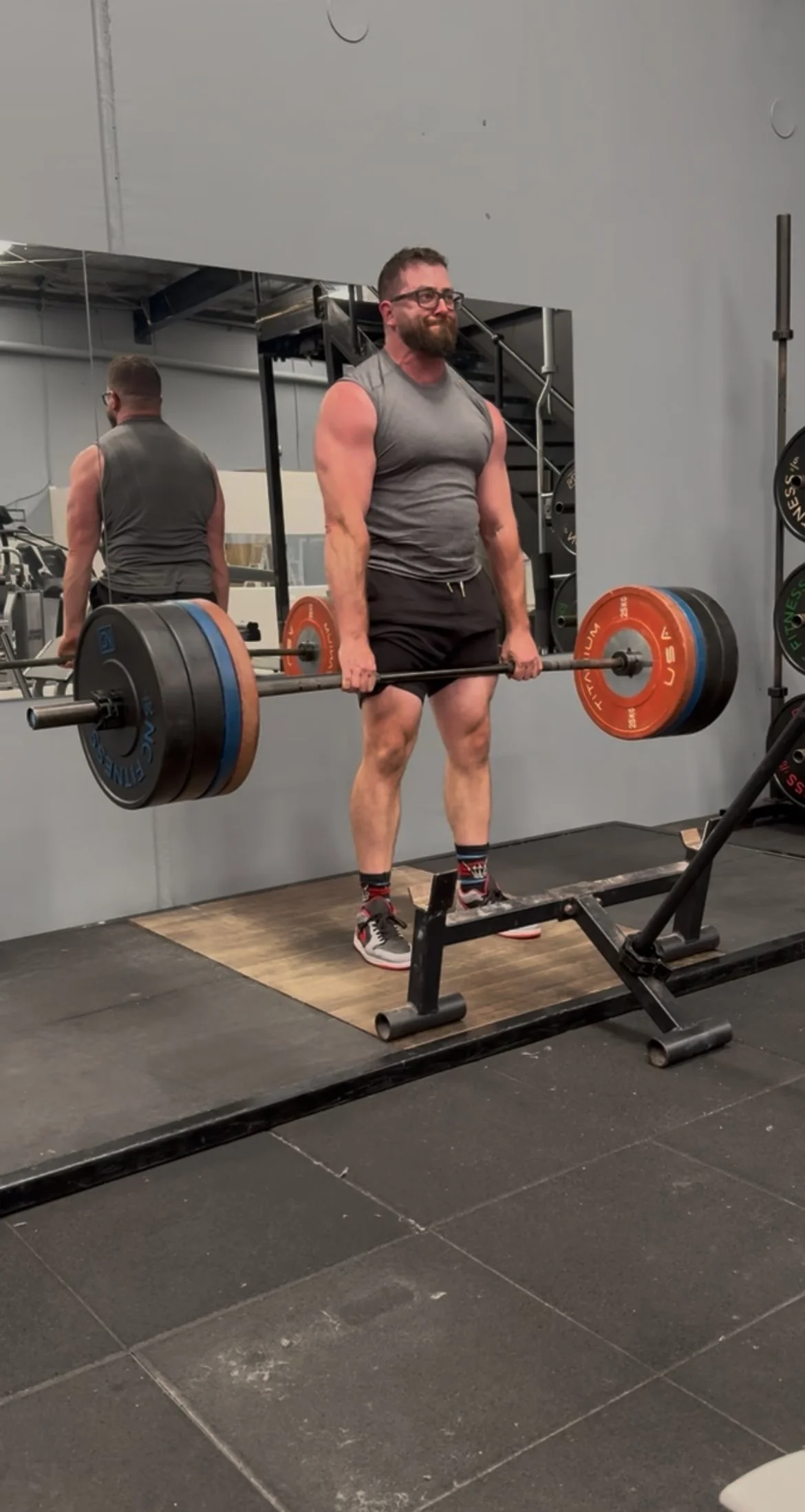 A man lifting a heavy barbell with weights in a gym, standing on a platform, reflected in a mirror.