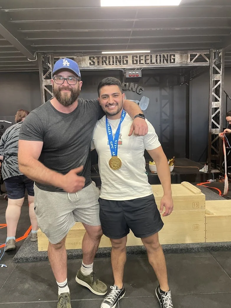 Two men posing together in a gym. One man is wearing a blue LA cap, glasses, a gray t-shirt, and light shorts. The other man is wearing a white t-shirt, black shorts, and has a gold medal around his neck. The background has a sign that reads 'Strong 