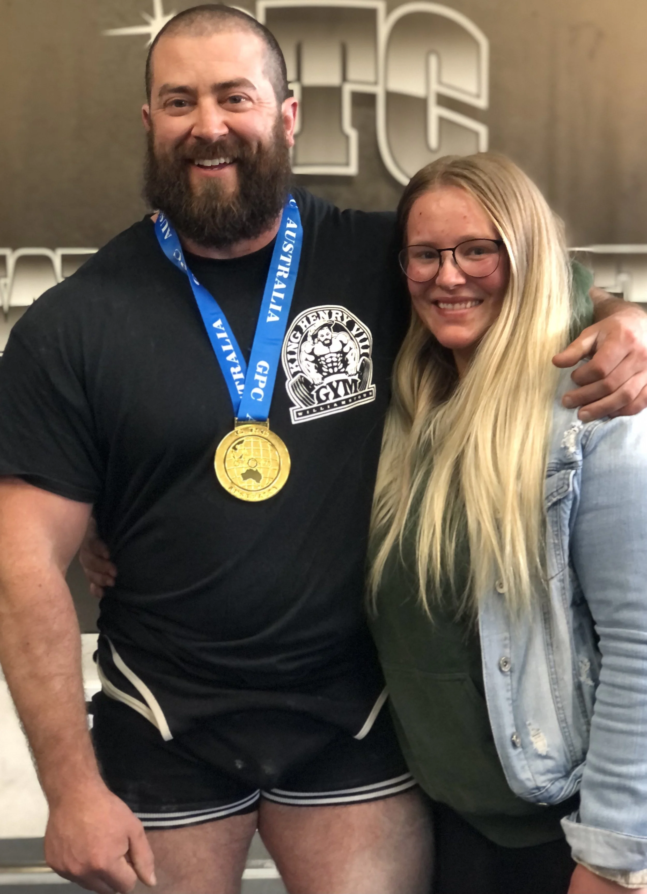 A muscular man with a beard, wearing a black T-shirt with a gym logo, black shorts with white trim, and a gold medal around his neck, standing next to a smiling woman with long blonde hair and glasses, in a gym setting.