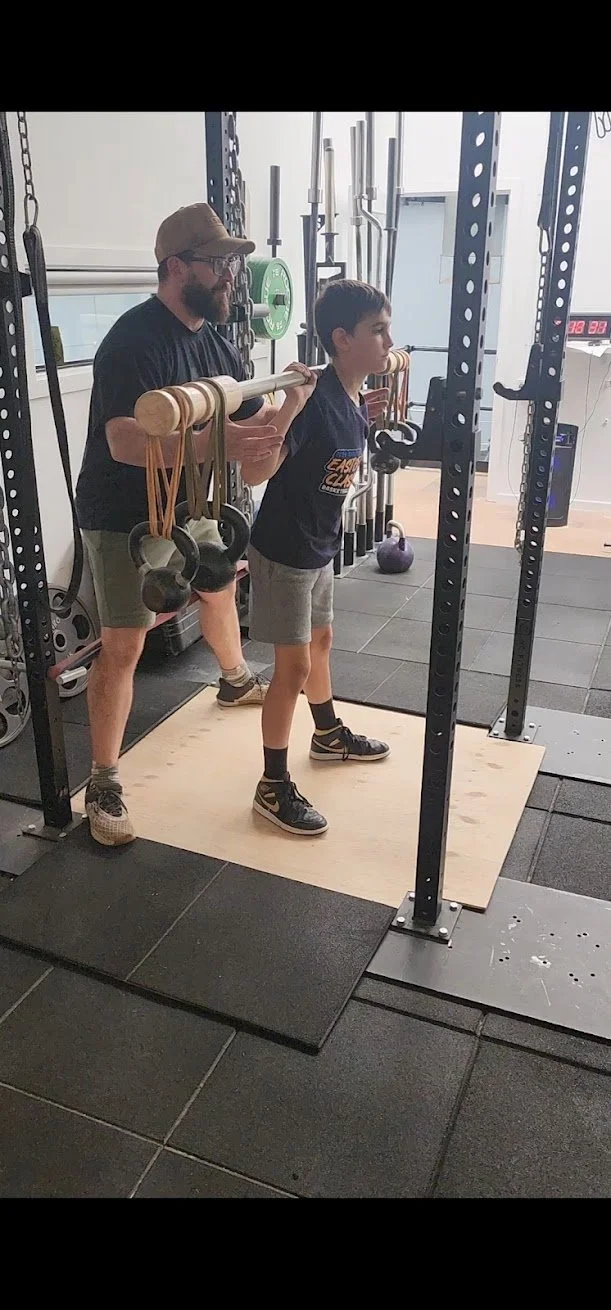 A young boy is performing a squat exercise in a gym with the help of an adult male trainer who is holding a wooden stick with gym equipment weights hanging from it behind his neck, providing guidance for proper form.