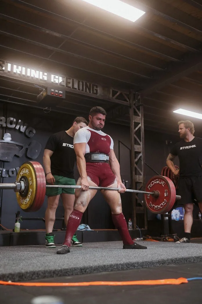 A powerlifter in red and white gear performing a deadlift at a competition, watched by two spotters in black.