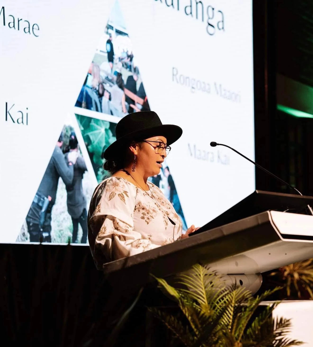 A woman wearing a floral blouse, black hat, and glasses giving a speech at a podium during a formal event with a large presentation screen behind her displaying text and photos.