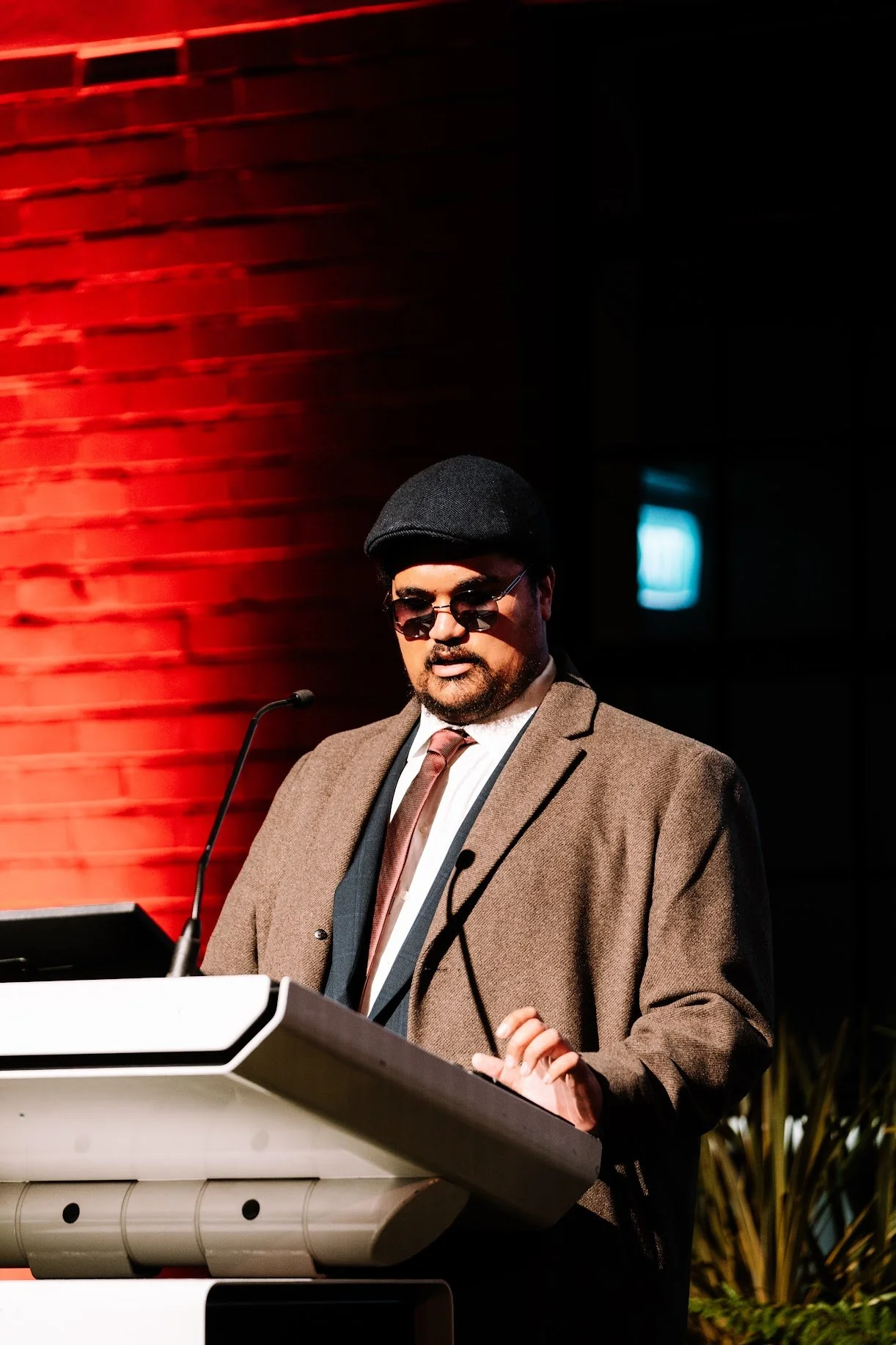 A man wearing a brown blazer, white shirt, tie, black hat, and sunglasses, standing at a podium with a microphone, in front of a red brick wall.