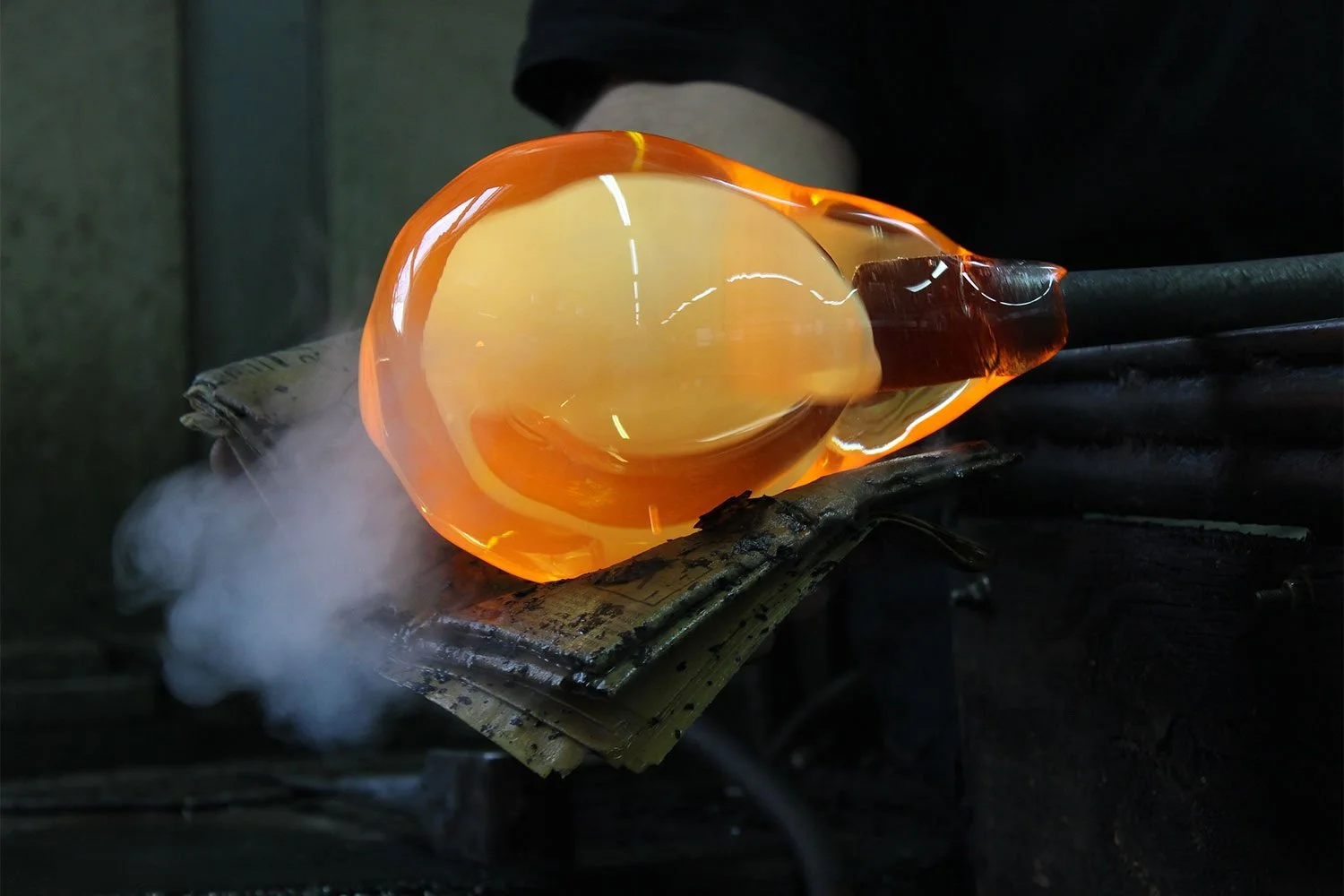 Glassblowing process with molten glass being shaped on a metal rod, with bright orange glow and smoke around.