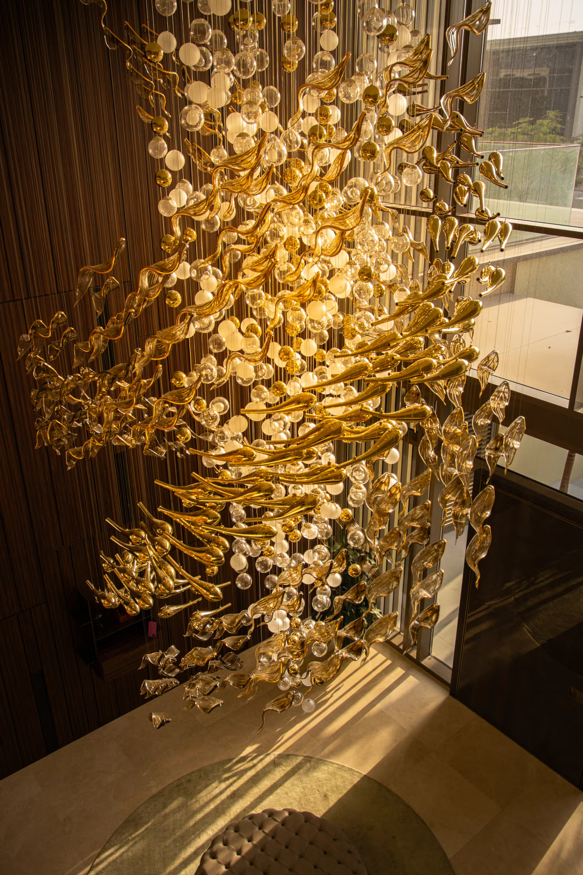 A modern chandelier made of glass and gold-colored metal swirls hanging inside a building near a large window.