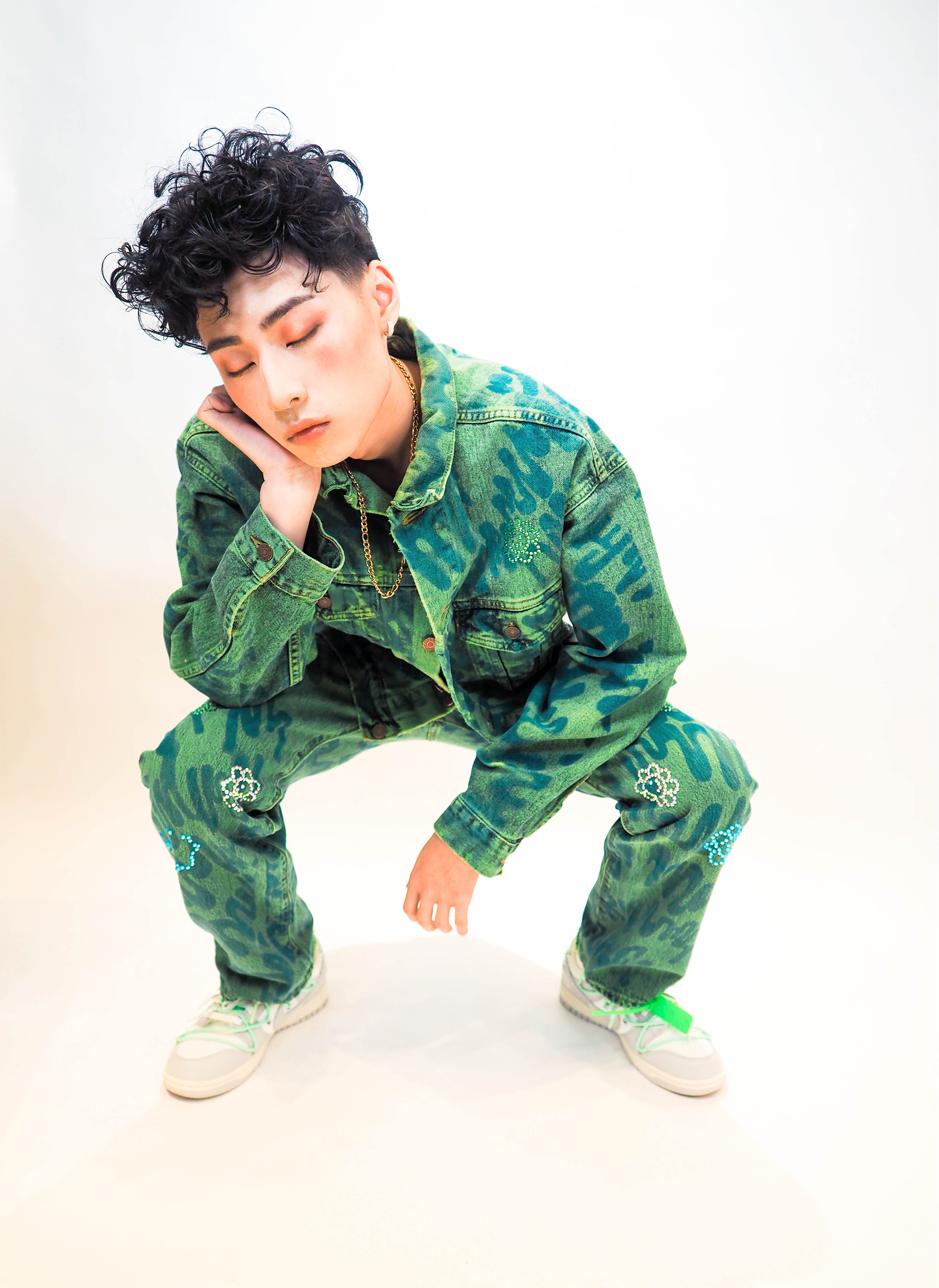 Person crouching in a squatting position wearing a green camouflage jacket and pants with decorative embellishments, white sneakers with green accents, and a gold chain necklace, with eyes closed and one hand resting on their face.