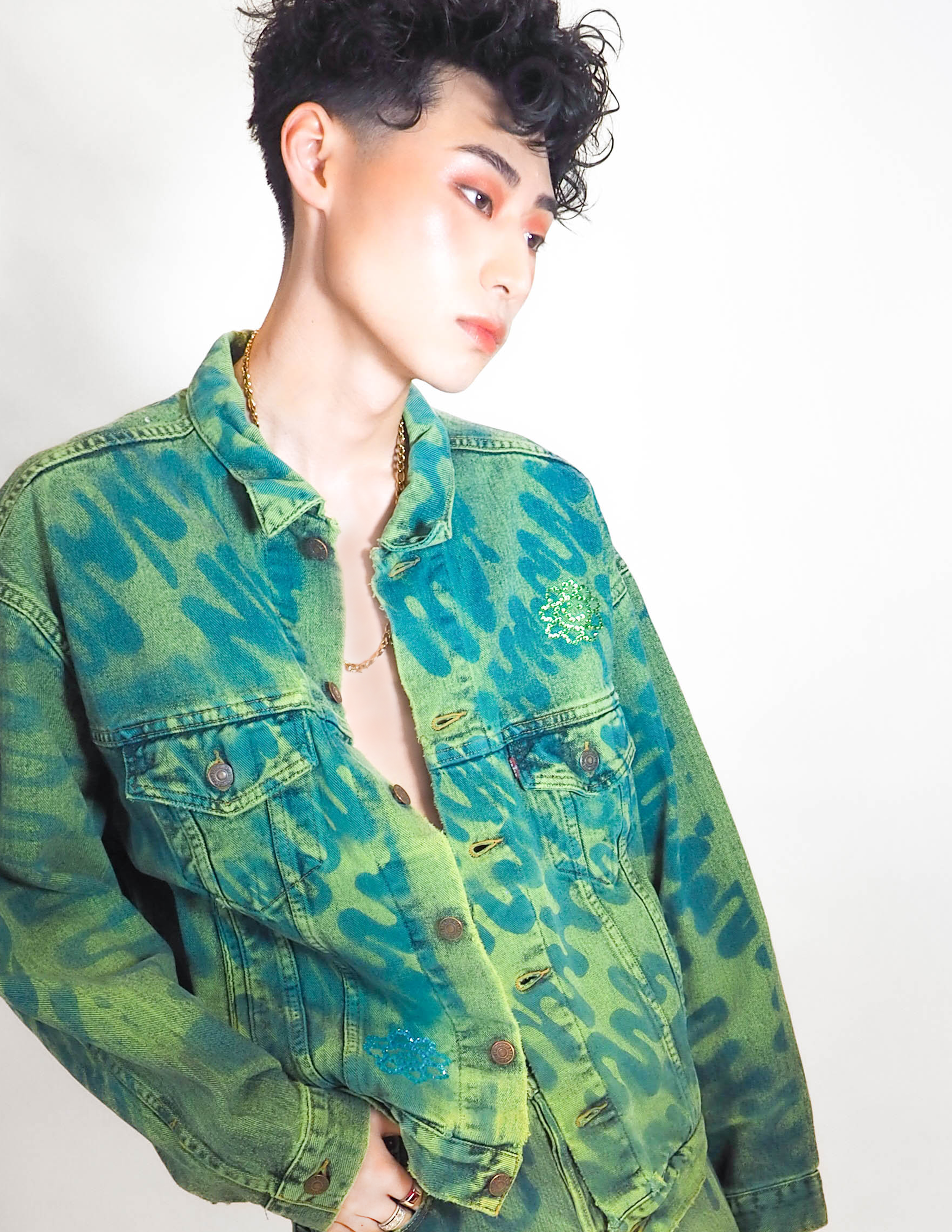 A person with short, curly black hair and makeup wearing a green-dyed denim jacket with blue abstract patterns and patch embellishments, standing against a plain background.