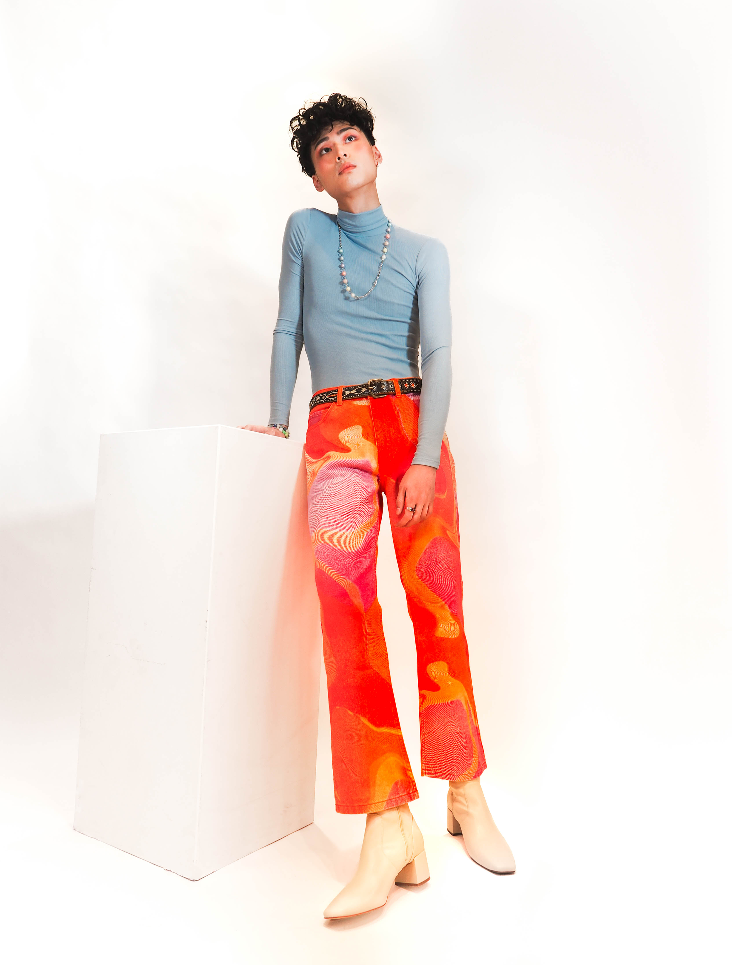 A person with short curly hair wearing a light blue turtleneck, colorful patterned pants, and cream-colored boots, standing next to a white pedestal against a plain white background.