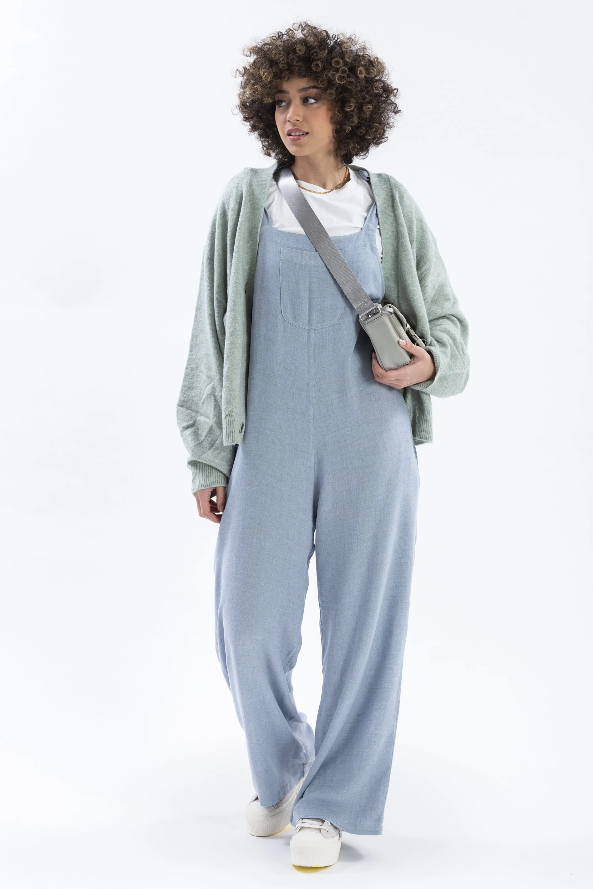 Young woman with curly hair in casual outfit, wearing a light green cardigan, white T-shirt, blue overalls, white sneakers, and carrying a gray shoulder bag, standing against a plain white background.