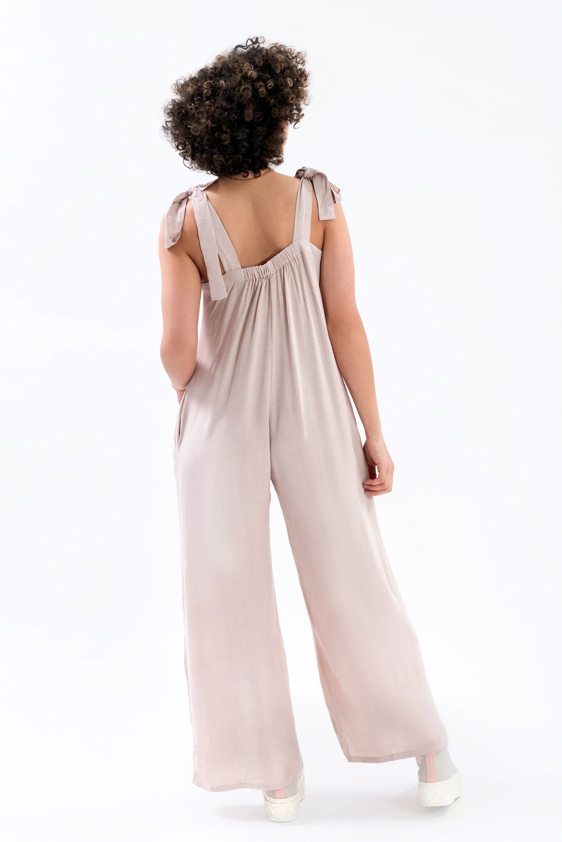 Back view of a woman with curly hair wearing a taupe-colored jumpsuit with tied shoulder straps and wide-leg pants, standing against a plain white background.