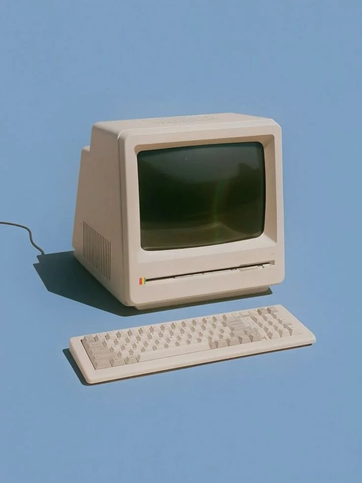 An old beige Apple Macintosh computer with a CRT monitor, keyboard, and a small rainbow-colored strip below the screen, set against a blue background.