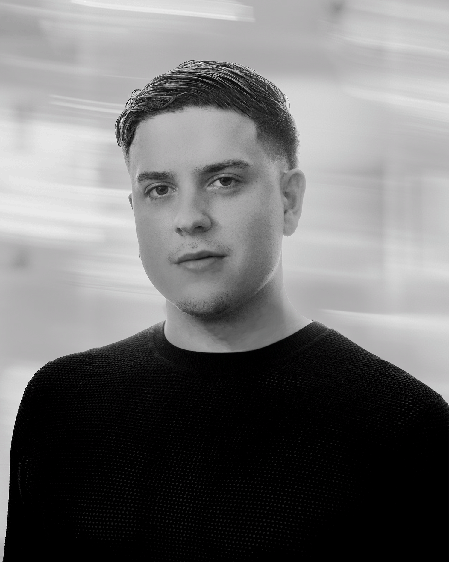 Black and white portrait of a young man with short hair, wearing a dark sweater, looking at the camera, with a blurred background.