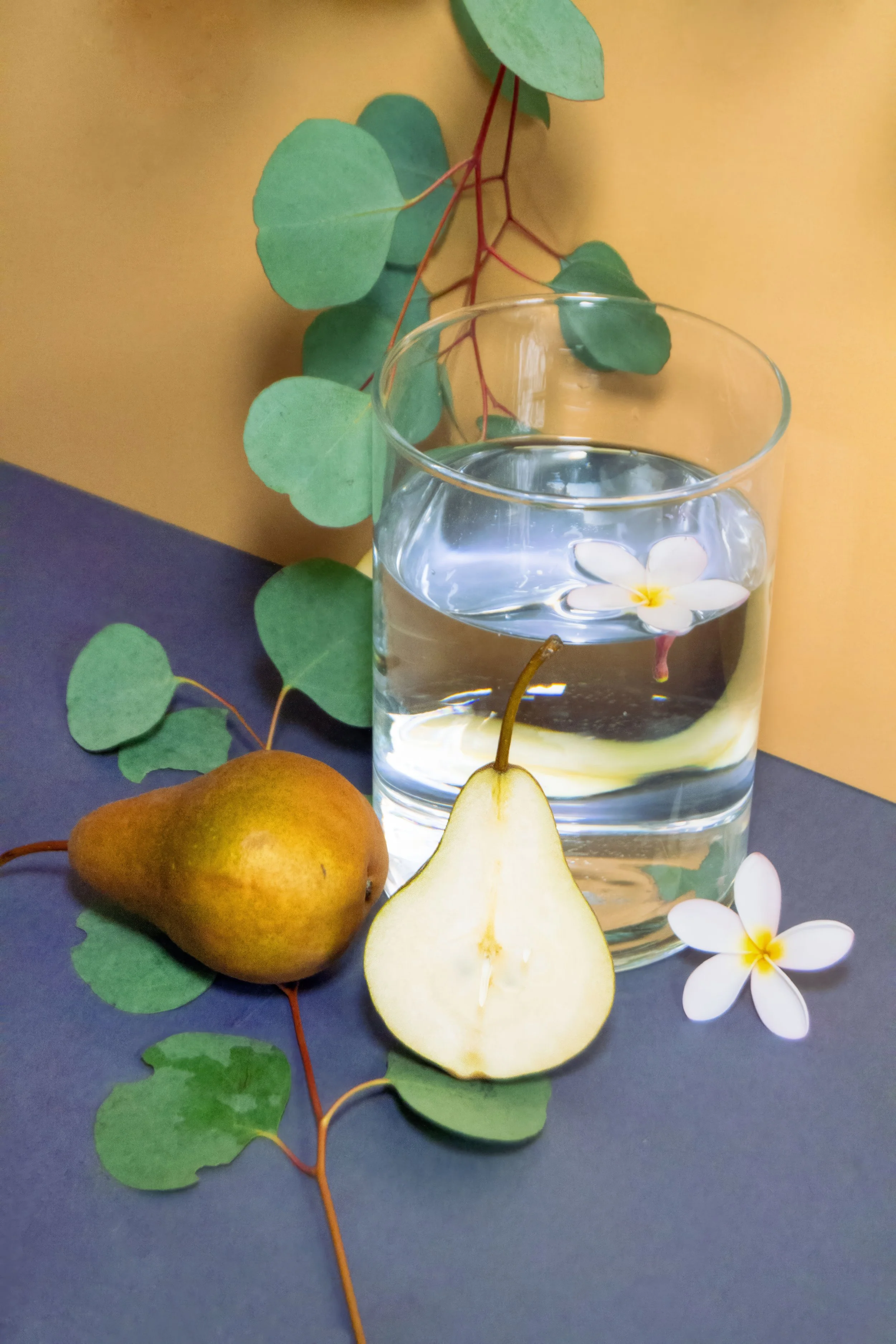 A glass of water with a floating flower, a pear, a half-cut pear, green leafy vines, and white flowers on a dark surface against a yellow background.