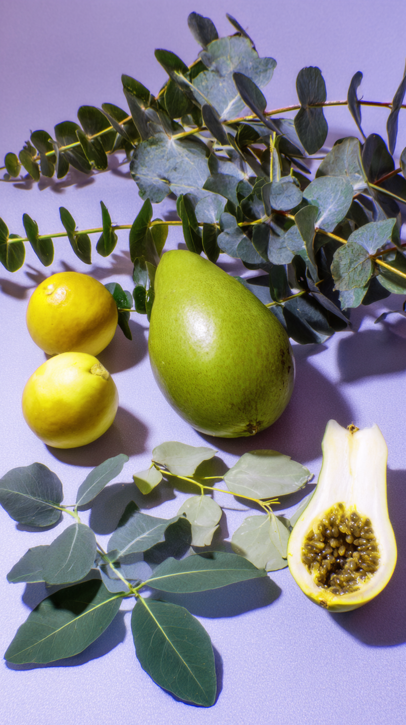 A still life photo of two yellow apples, one green pear, two papayas with seeds visible, and eucalyptus leaves on a pastel purple background.