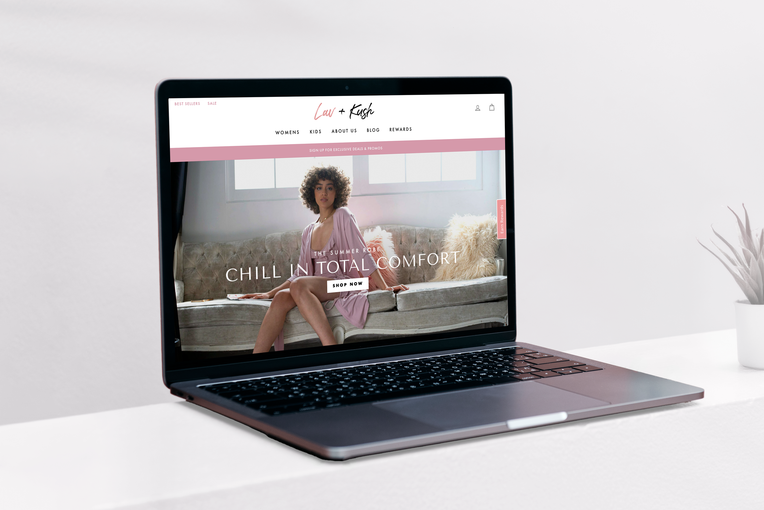 Open laptop on a white table displaying an online clothing store featuring a woman sitting on a beige sofa in pink loungewear, with website menus and promotional banner.