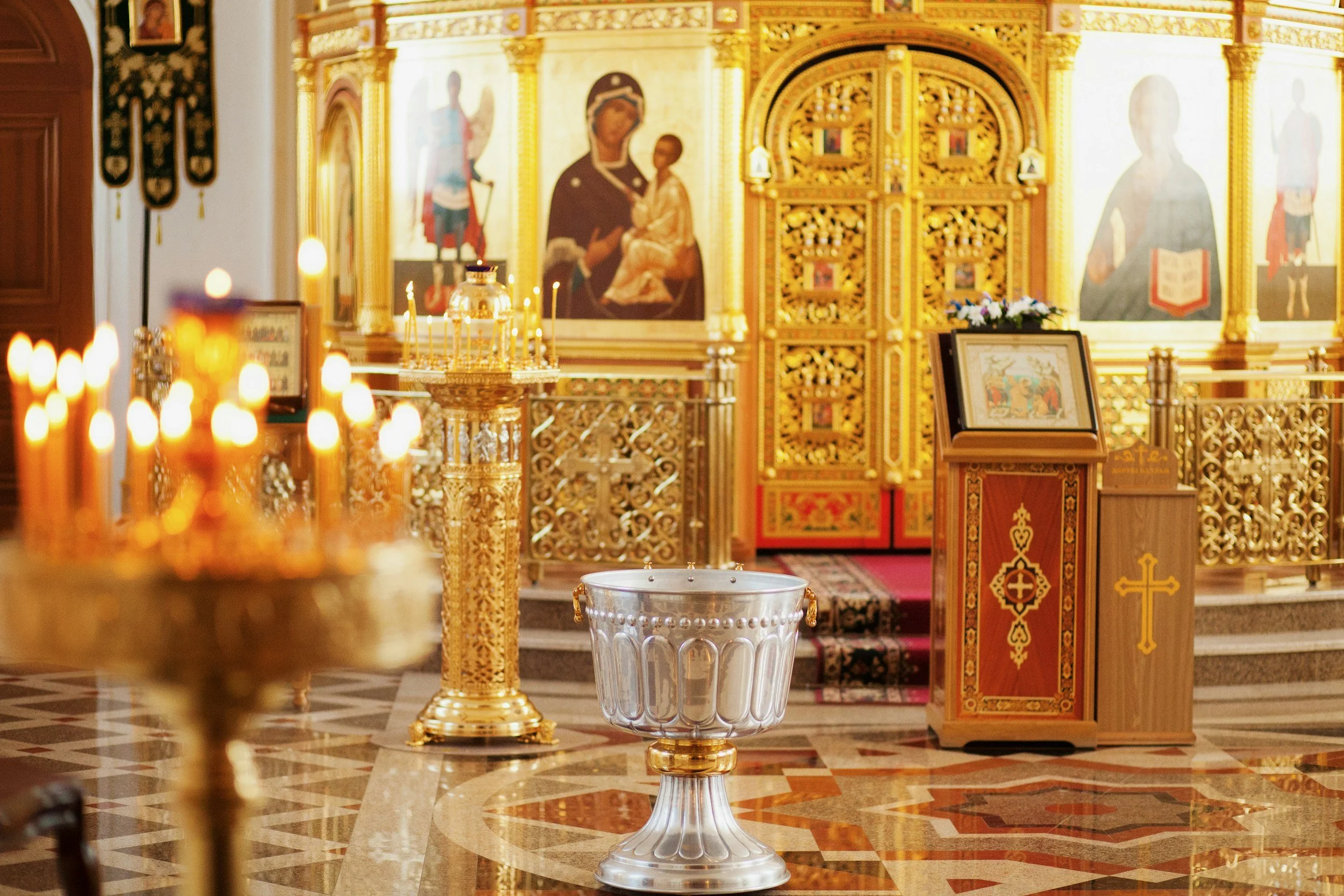 Inside an ornate church or cathedral with golden decorations, religious icons, and icons of saints on the walls, candleholders with lit candles, a baptismal font in the foreground, and an altar at the back with icons and flowers.