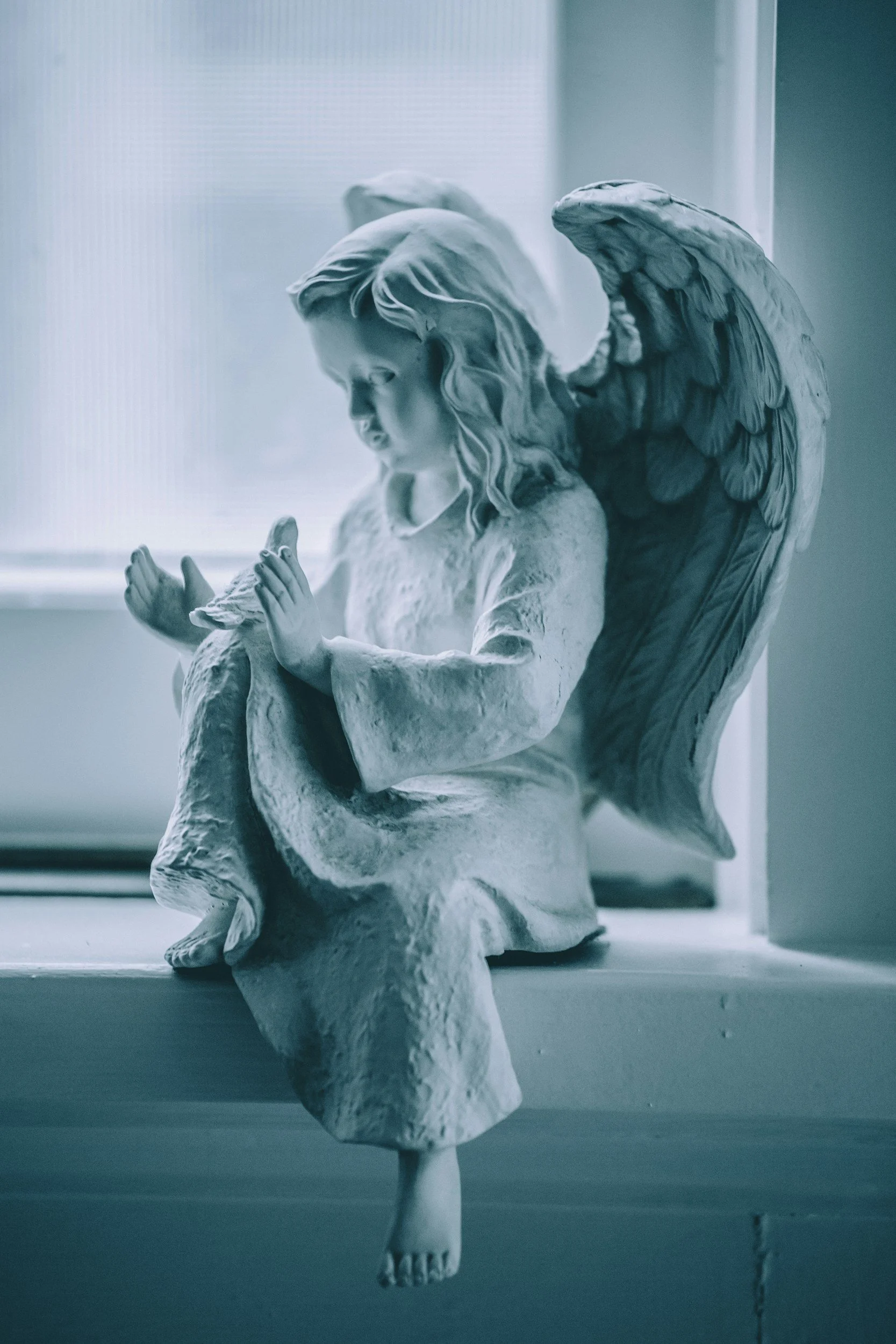 A white angel figurine with detailed wings kneeling on a windowsill with hands clasped.