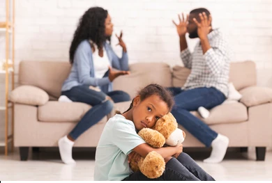 Child sitting on floor hugging teddy bear while two adults argue on sofa in background.
