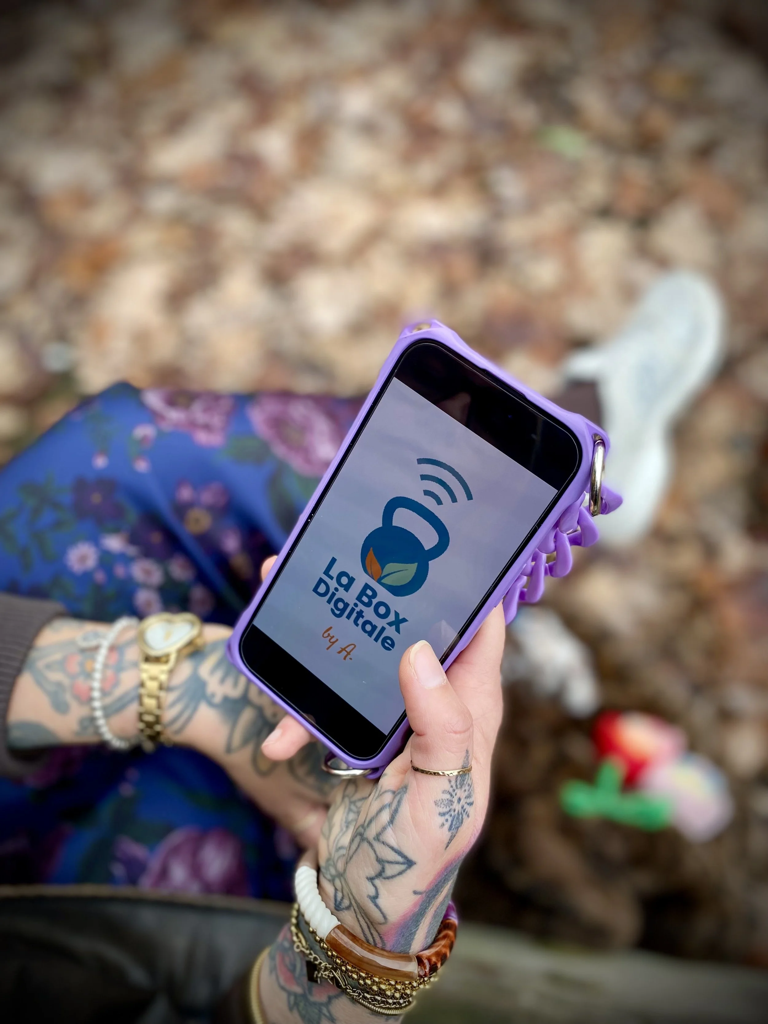 Une personne tenant un téléphone portable avec l'écran affichant le logo 'La Box Digitale'. La personne porte plusieurs bracelets et a des tatouages sur la main. En arrière-plan, il y a des feuilles mortes sur le sol.