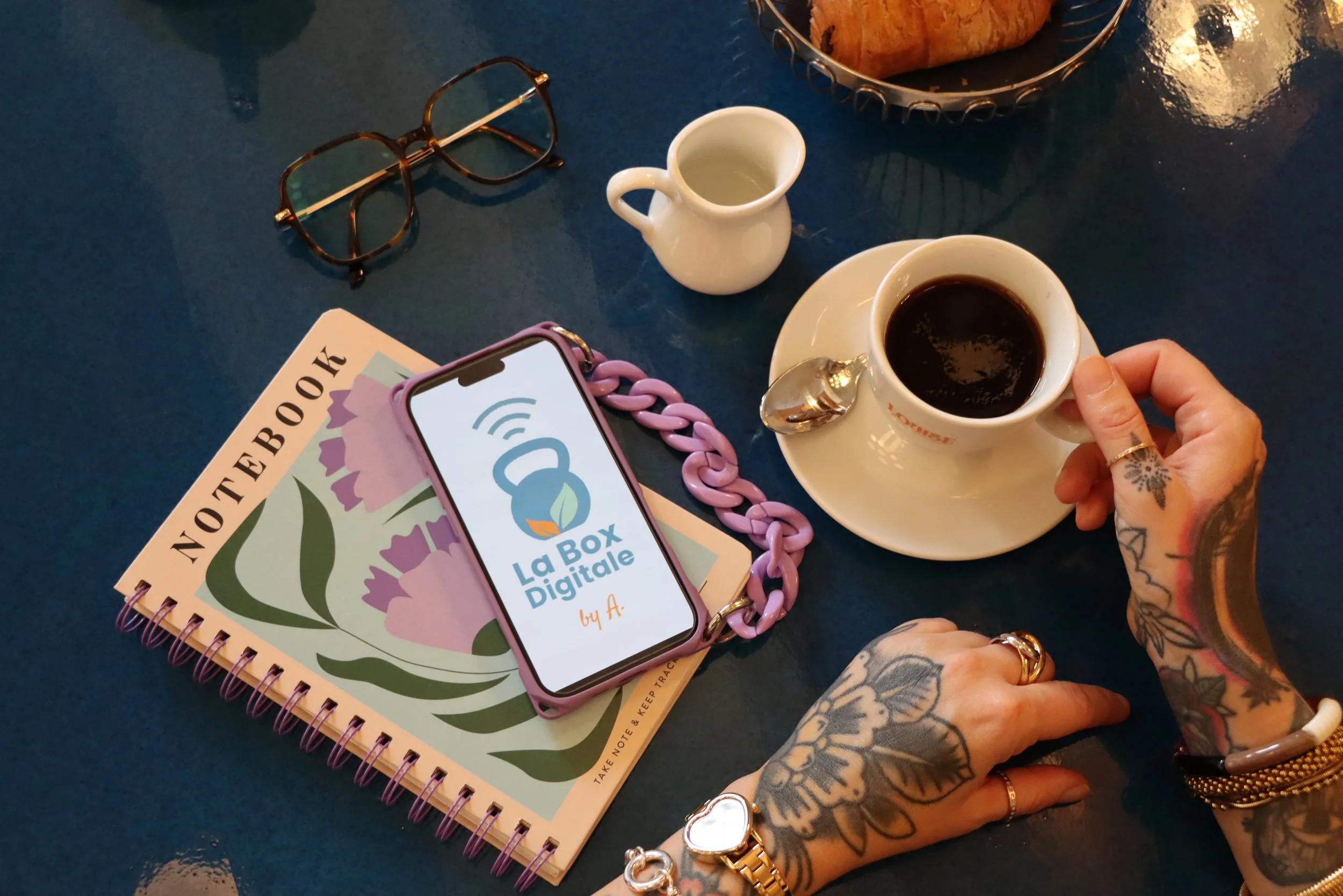 Une personne aux mains tatouées tenant une tasse de café noir sur une table. Sur la table, il y a aussi une autre tasse vide, un carnet avec la mention 'NOTEBOOK' et une tablette avec un logo 'La Box Digitale'.