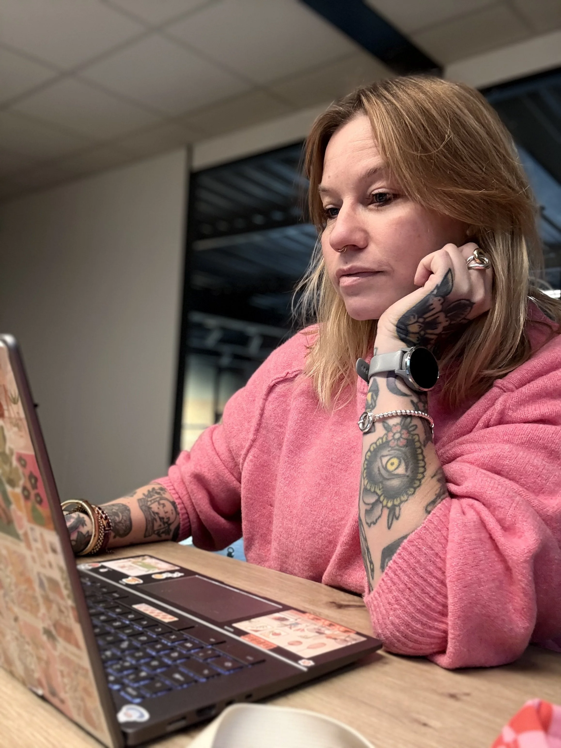 Une femme aux tatouages, portant un bracelet et une montre, regarde un ordinateur portable en café ou espace de travail.
