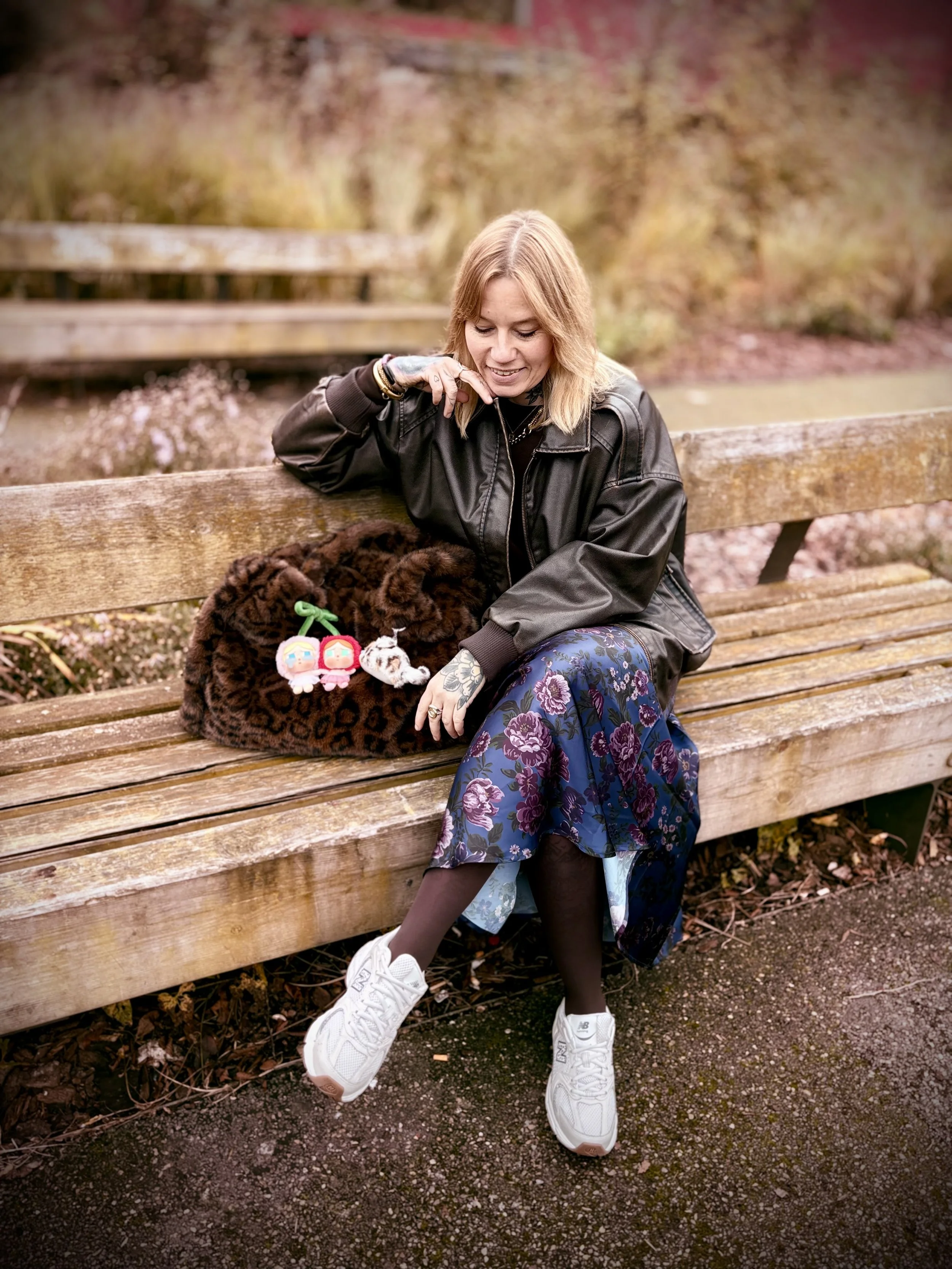Jeune femme assise sur un banc en bois dans un parc, portant une jupe longue à fleurs, un blouson en cuir, des baskets blanches, avec un sac à motif léopard. Devant elle, un sac à main avec des porte-clés colorés. Elle sourit et regarde vers le bas, avec un fond de végétation automnale.