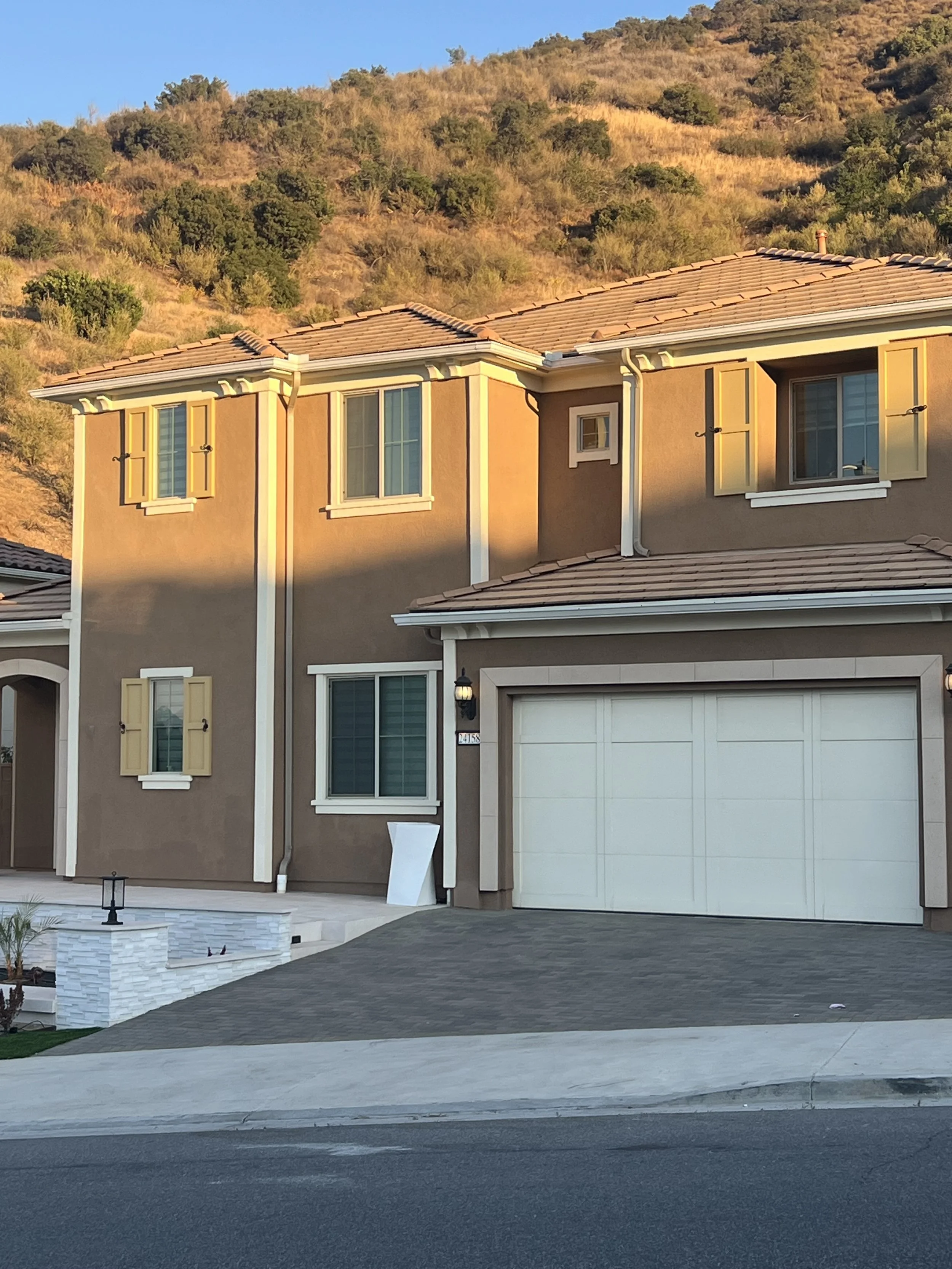 This is an image of a modern house with a garage, located in a hilly area with dry and green vegetation in the background. The house has beige stucco walls, white trim, yellow shutters, and a tiled roof. A small white structure is near the garage, an