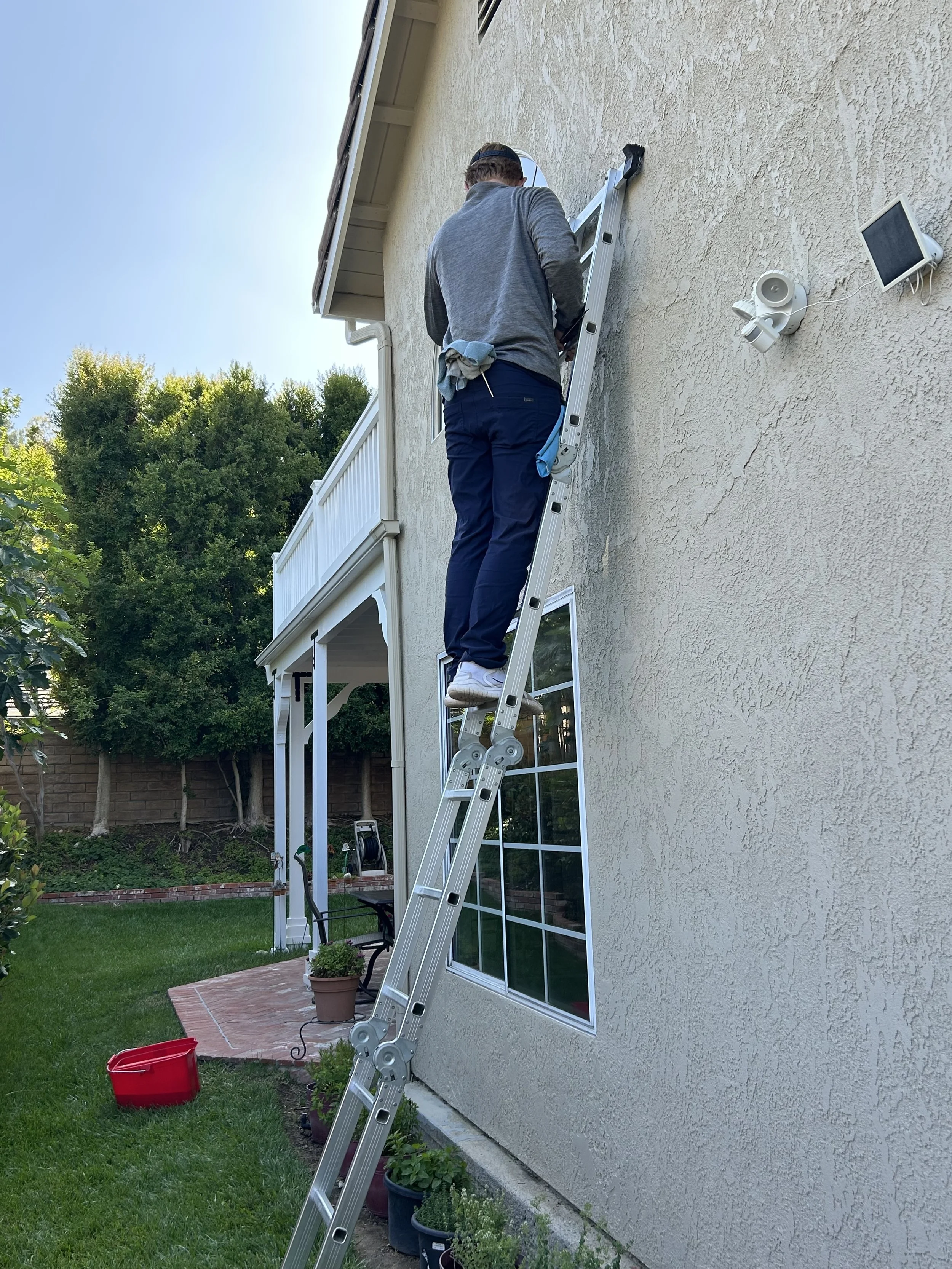 A man is standing on a ladder outside a house, working on the wall near a window. There are potted plants and a red bucket on the grass below.