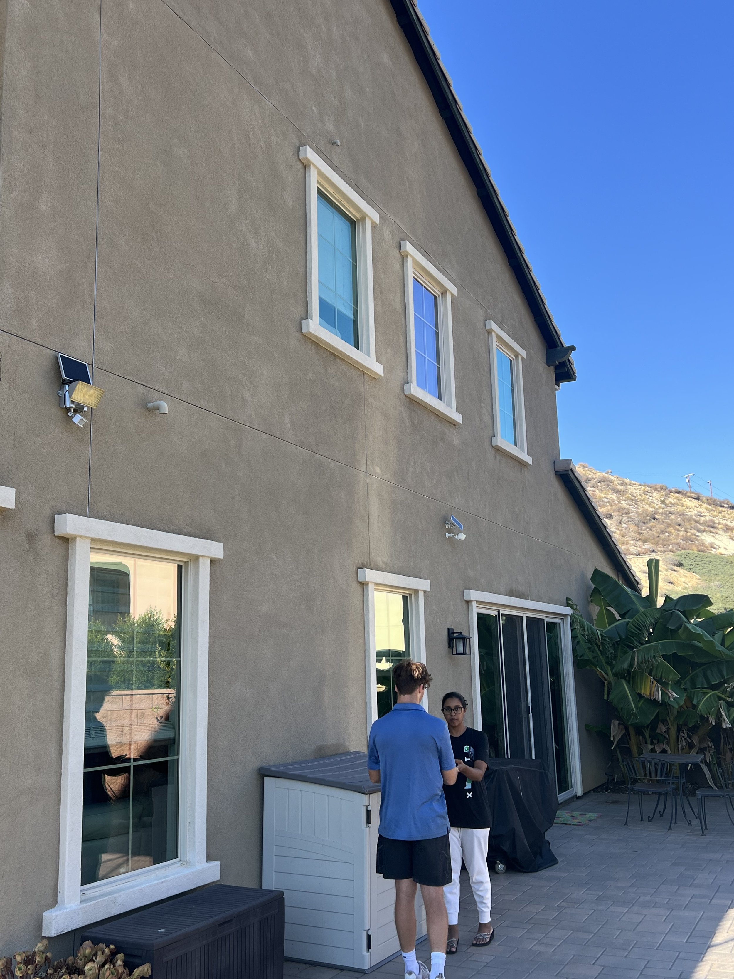 People standing in a backyard patio near a beige house with multiple white-trimmed windows, a black patio table, and lush green plants, with hills in the background on a sunny day.