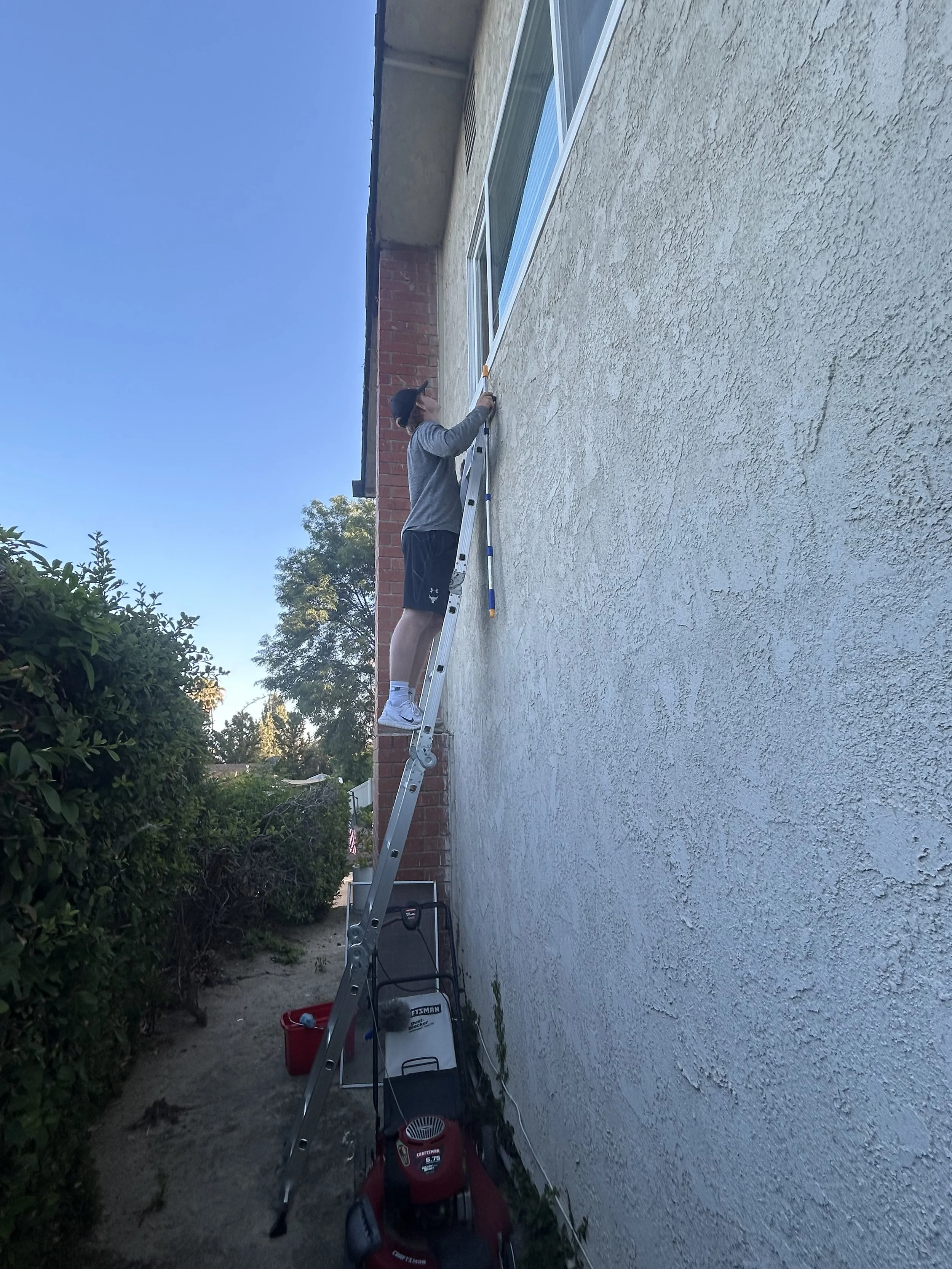 Person standing on a ladder, working on the exterior wall of a building near a window, with tools and equipment placed below.