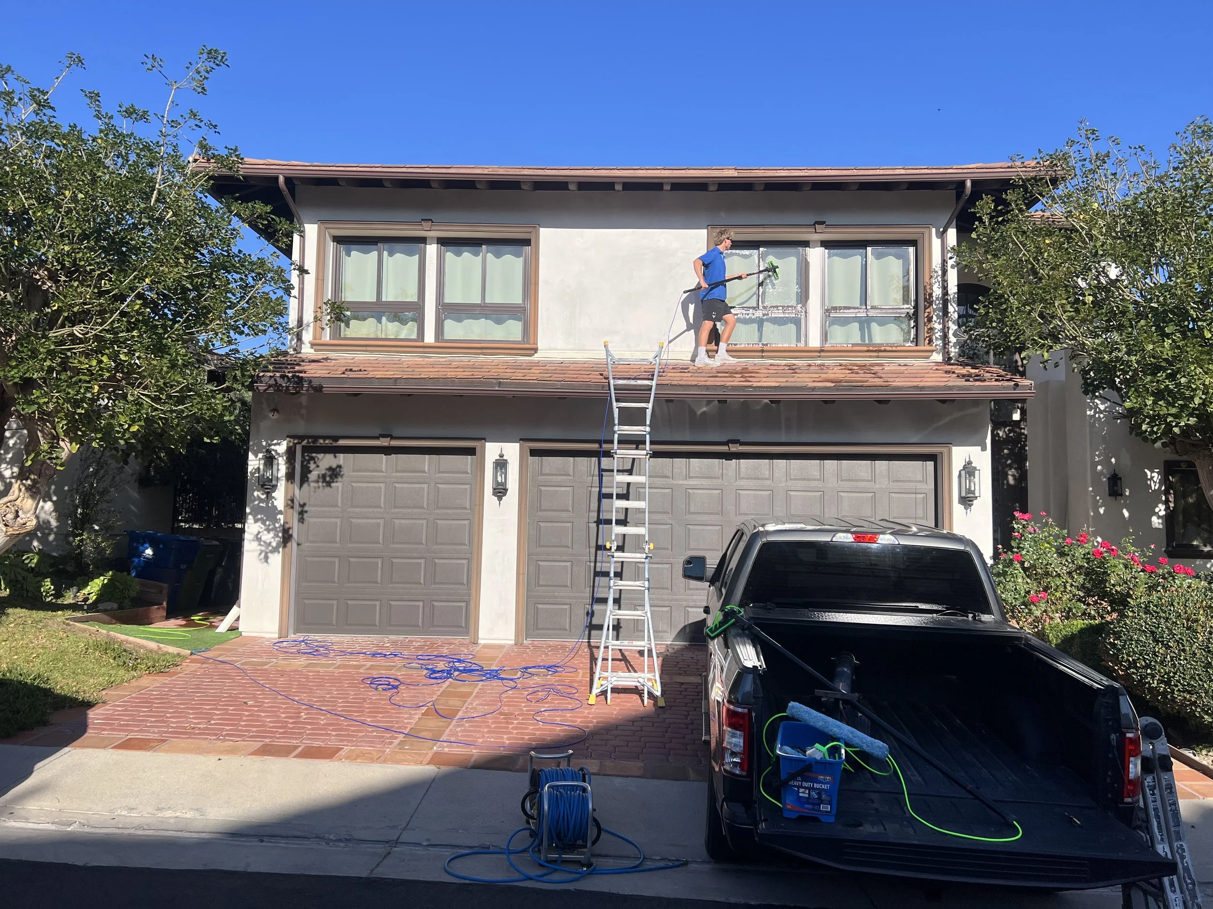 Person pressure washing the second story window of a house, with a ladder leaning against the house. Equipment and garden hoses are scattered on the driveway, and a car is parked in front.