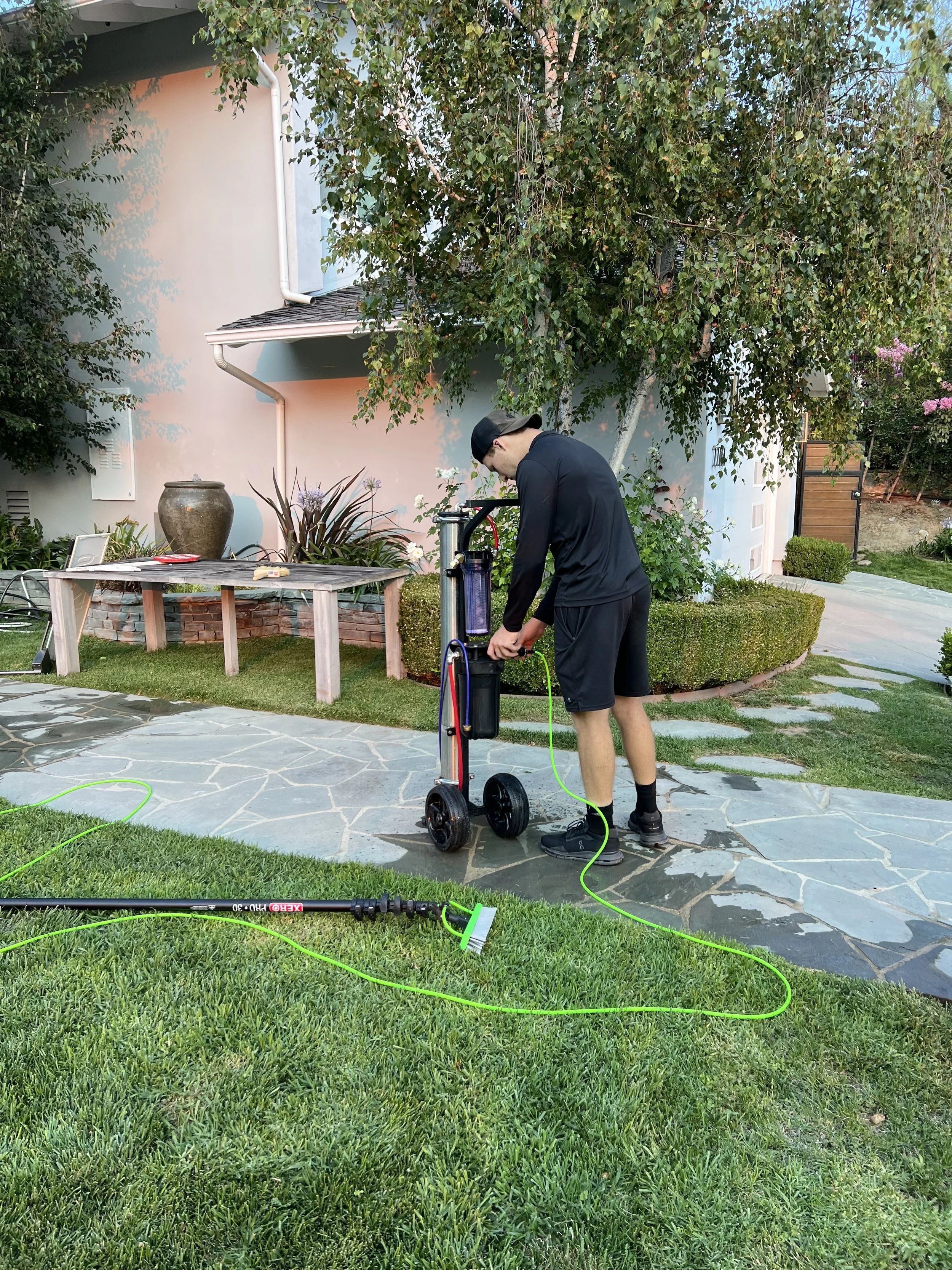 A man in black athletic clothes and a cap is outdoors on a stone patio, filling a water container with a water filtration system. There is a grassy yard with a large tree, a table with some items, and a house with a light-colored wall in the backgrou