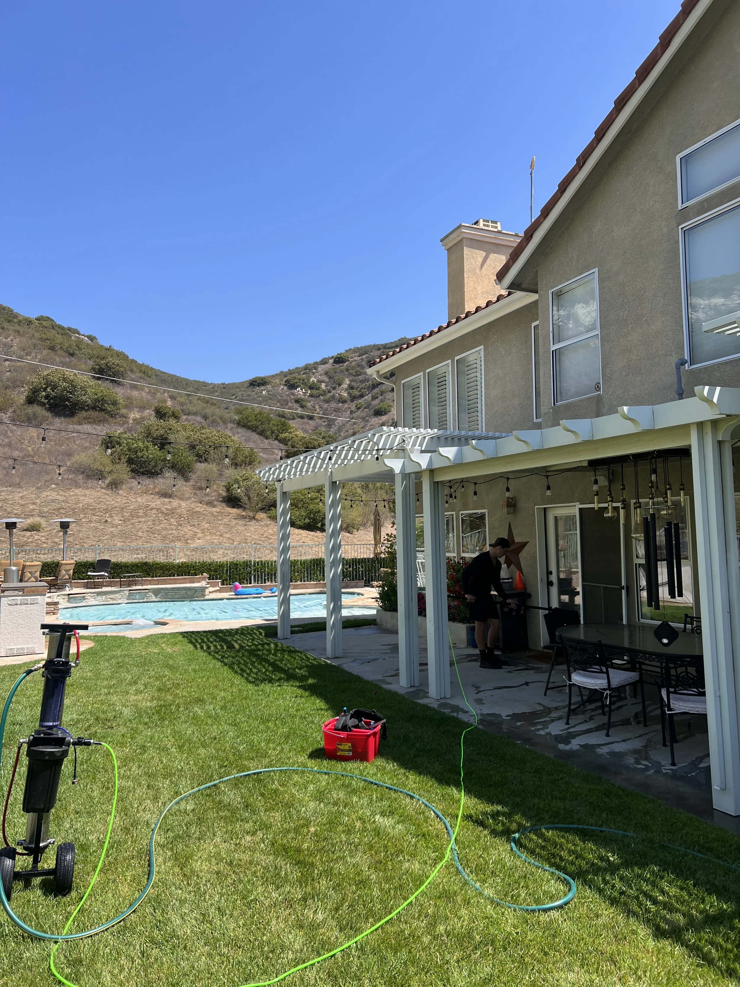 A backyard with a green lawn, a swimming pool, and a house with beige walls and a red tile roof. There are hills with bushes in the background. Items like a red container, outdoor furniture, and a person working outside can be seen, along with variou