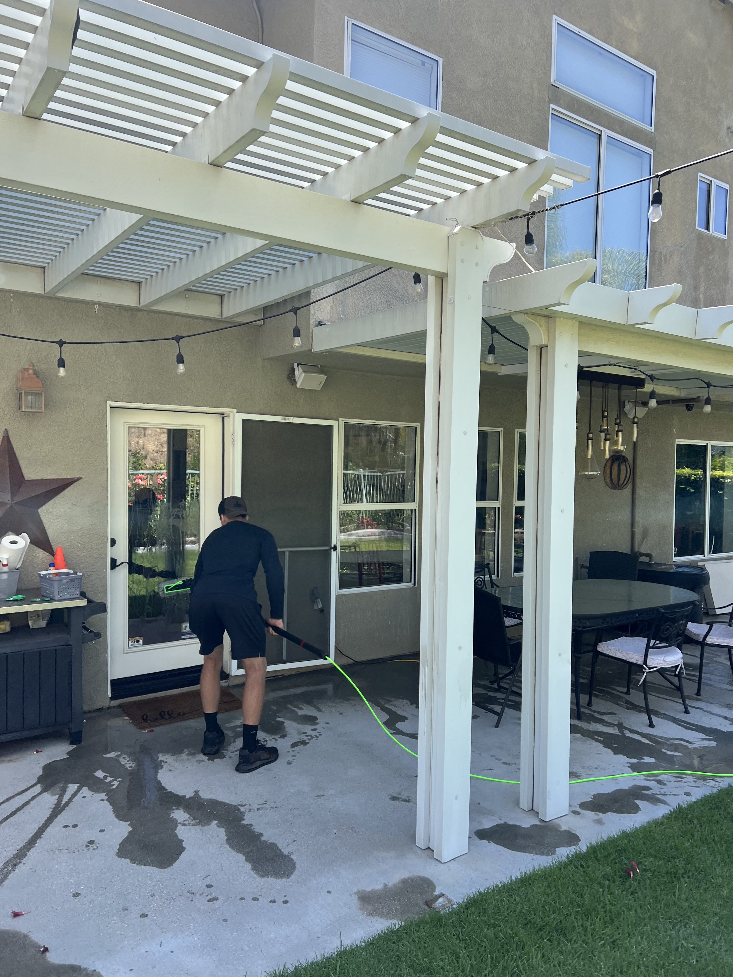 A man cleaning the patio floor outside a house. The house has large windows and a sliding glass door. There are outdoor string lights hanging, a table with chairs, and a star-shaped decoration on the wall.
