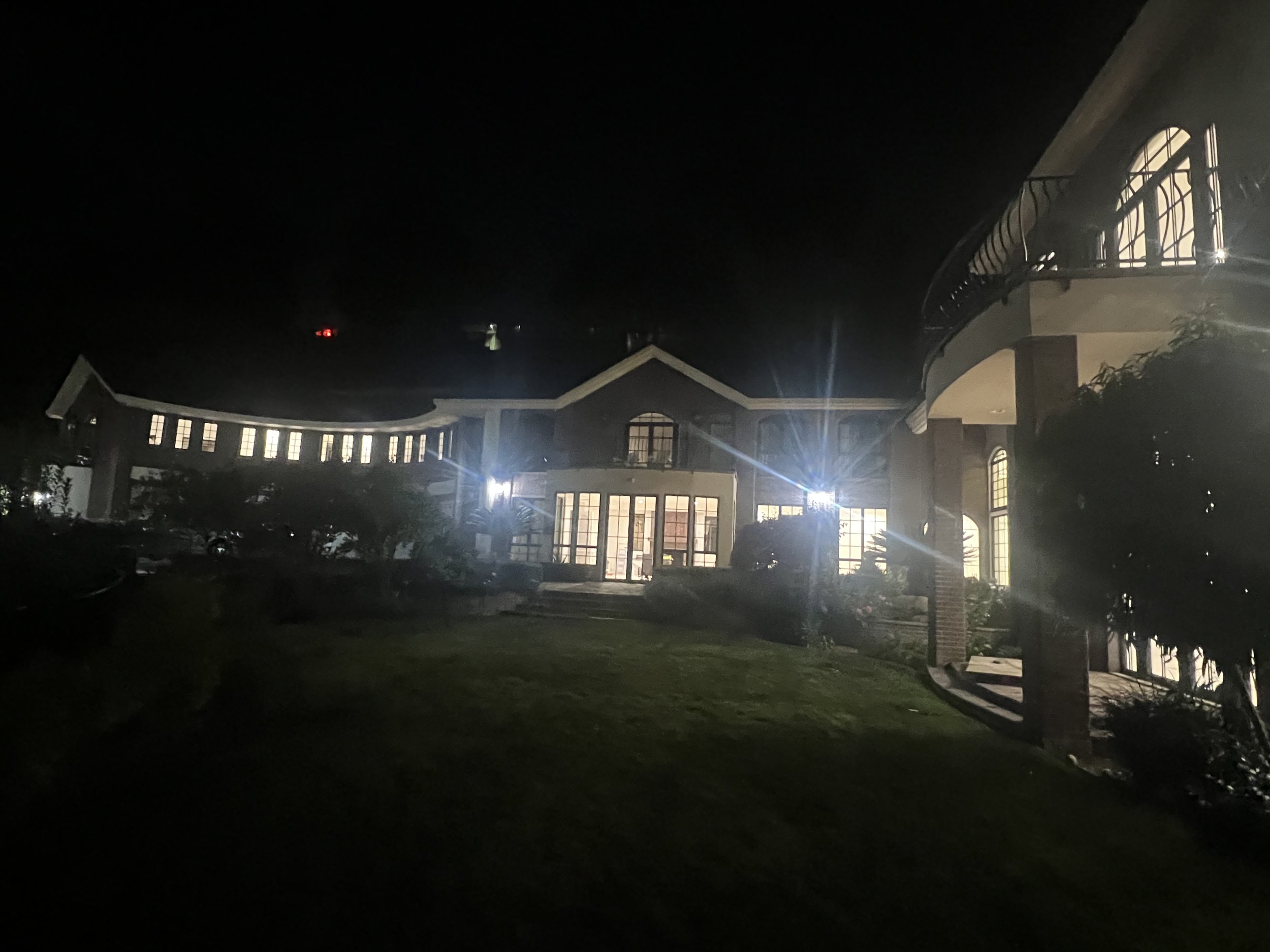 Nighttime view of a large house with many windows illuminated from within. The house features a curved balcony on the second floor and a well-maintained front yard with grass and bushes. Exterior lights are turned on, casting bright light onto parts 