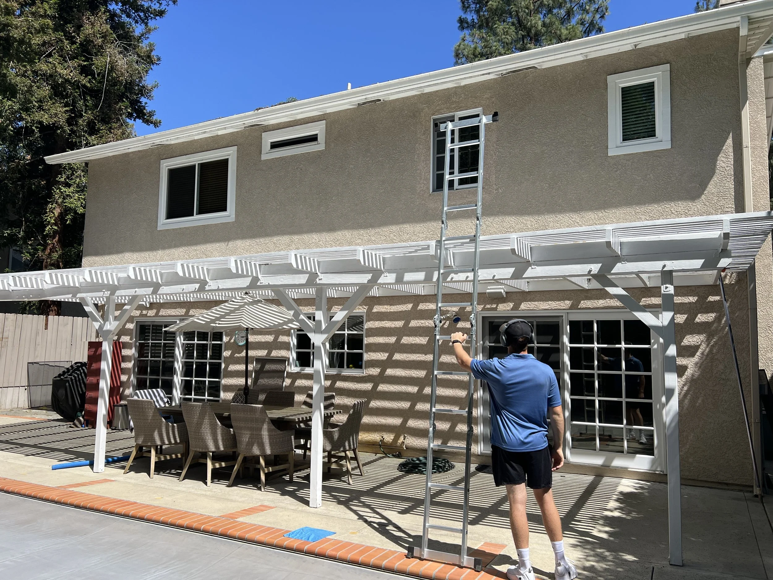 A person in a blue shirt and black shorts loading a ladder against the back of a beige house with white trim. There is a patio area with outdoor furniture and a white pergola casting shadows on the ground. The sky is blue and there are trees surround