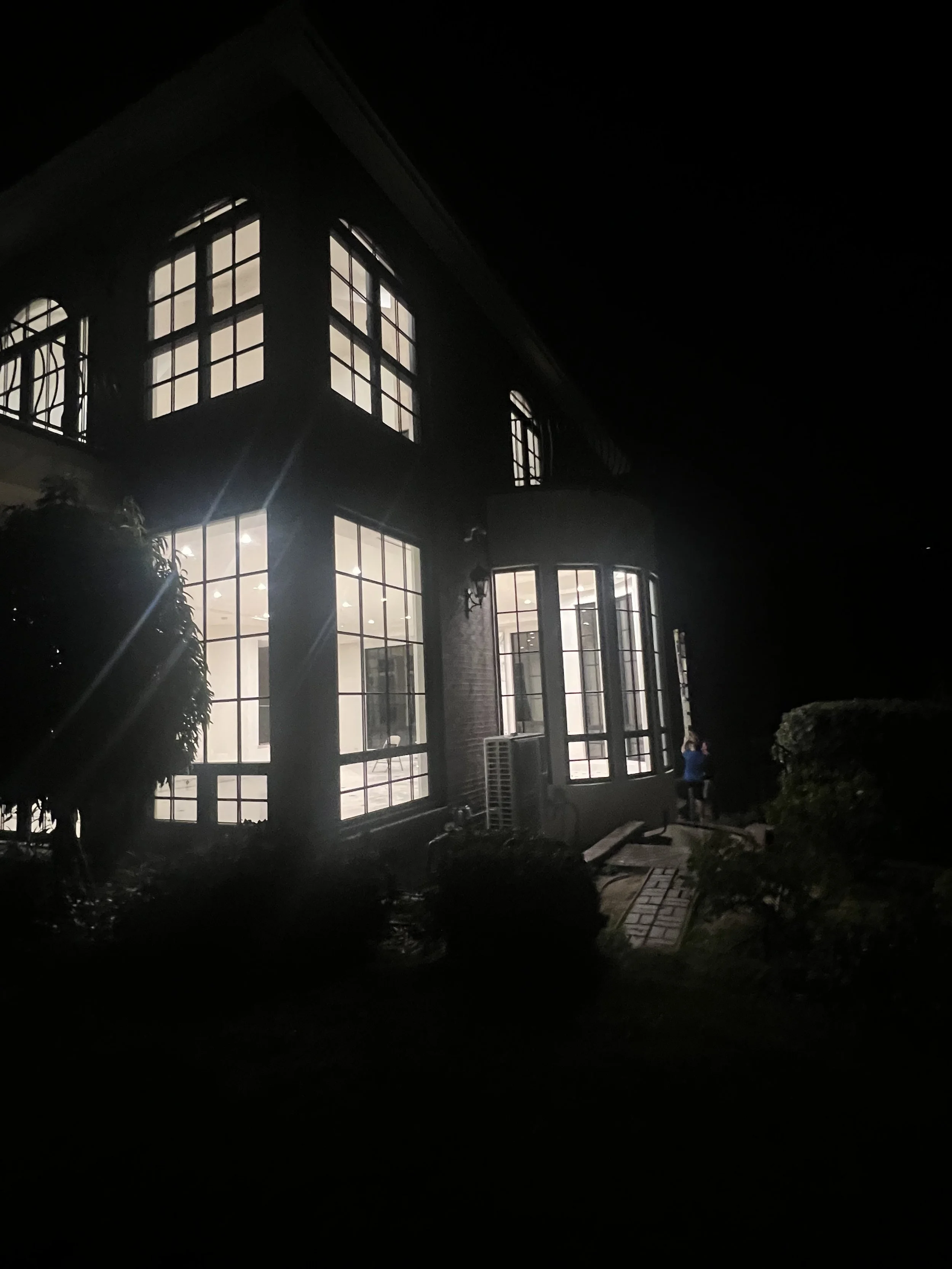 A large house with illuminated windows at night, featuring multiple large windows and a curved bay window, surrounded by a garden with plants and a paved walkway.
