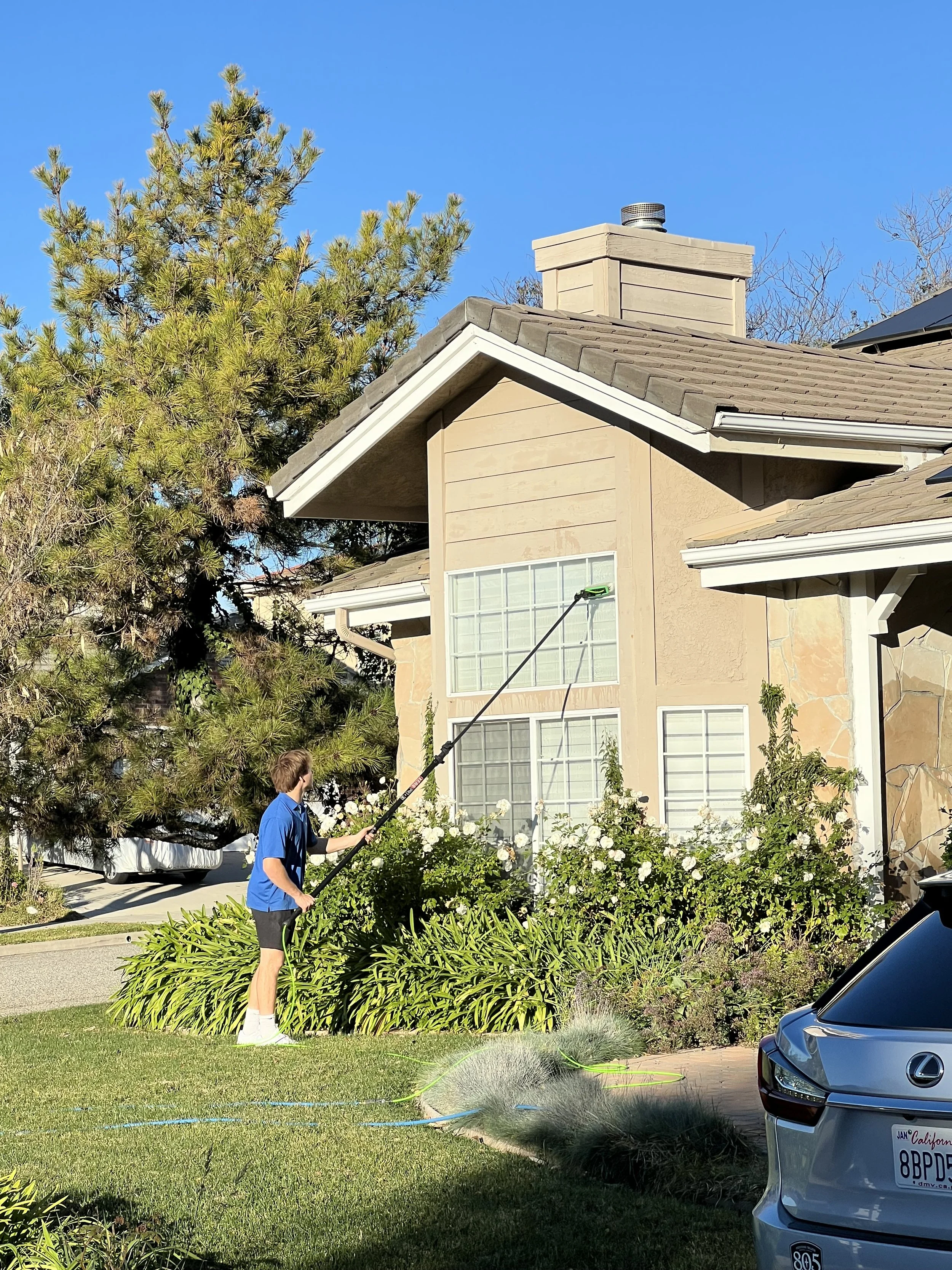 A young man in a blue shirt and black shorts is watering plants with a hose in a front yard. There is a beige house with white-framed windows, a stone pathway, and a green lawn with bushes and flowers. A car is parked in the driveway. The sky is clear and blue.