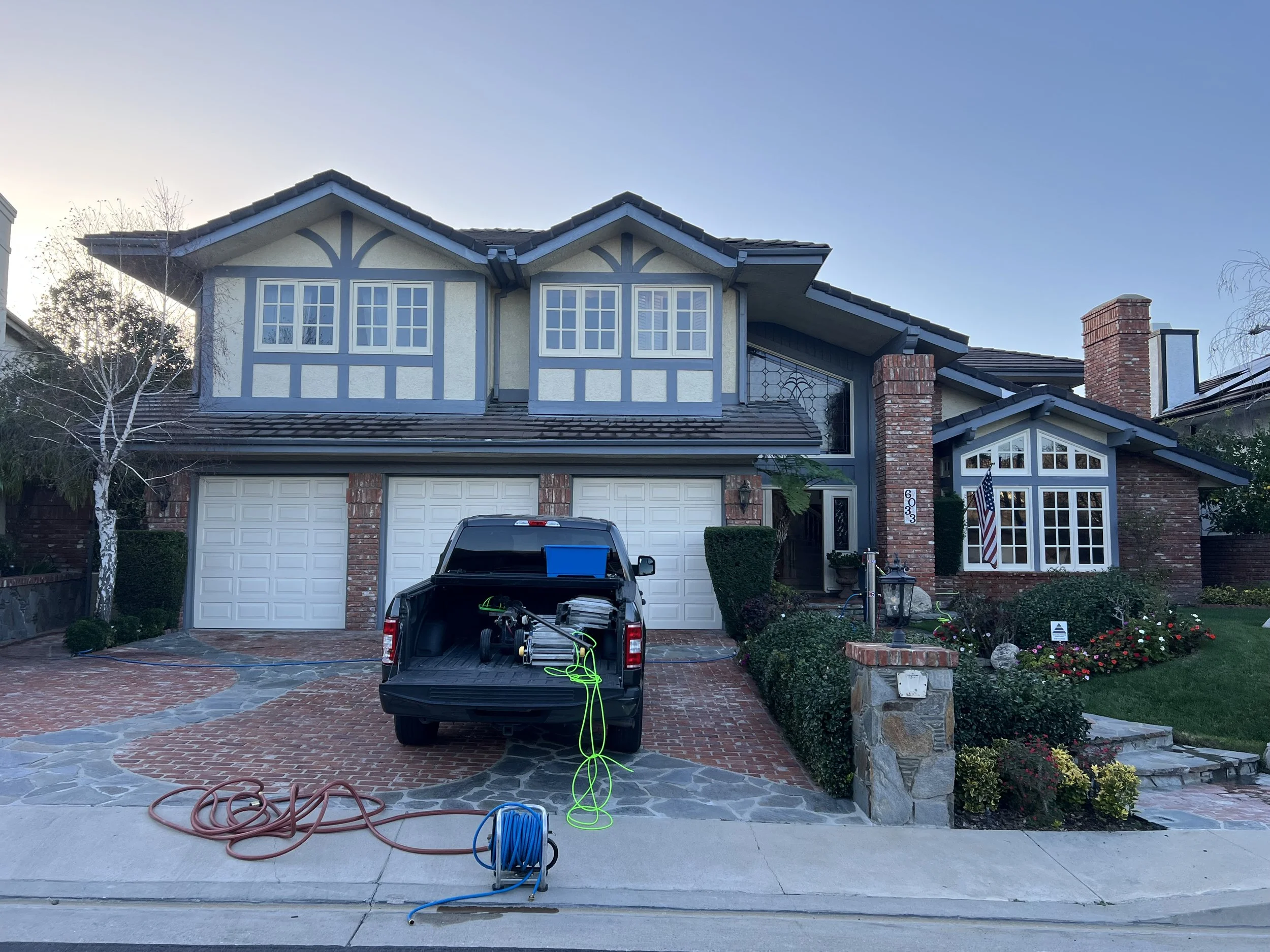Front view of a house with two garage doors, brick and blue siding exterior, and a brick walkway. There is a black pickup truck in front with garden hoses and extension cords. The house has multiple windows, some with decorative multiple panes, and a