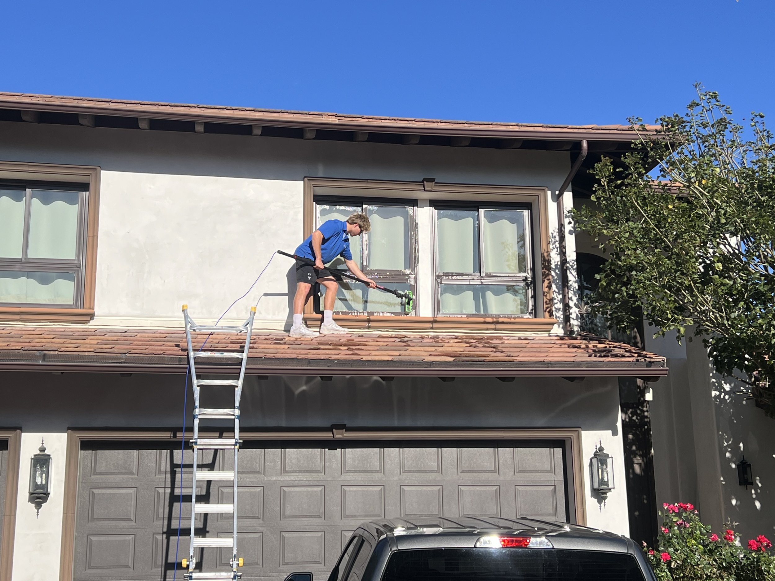 A person cleaning a second-story window of a house with a pressure washer, standing on a ladder.