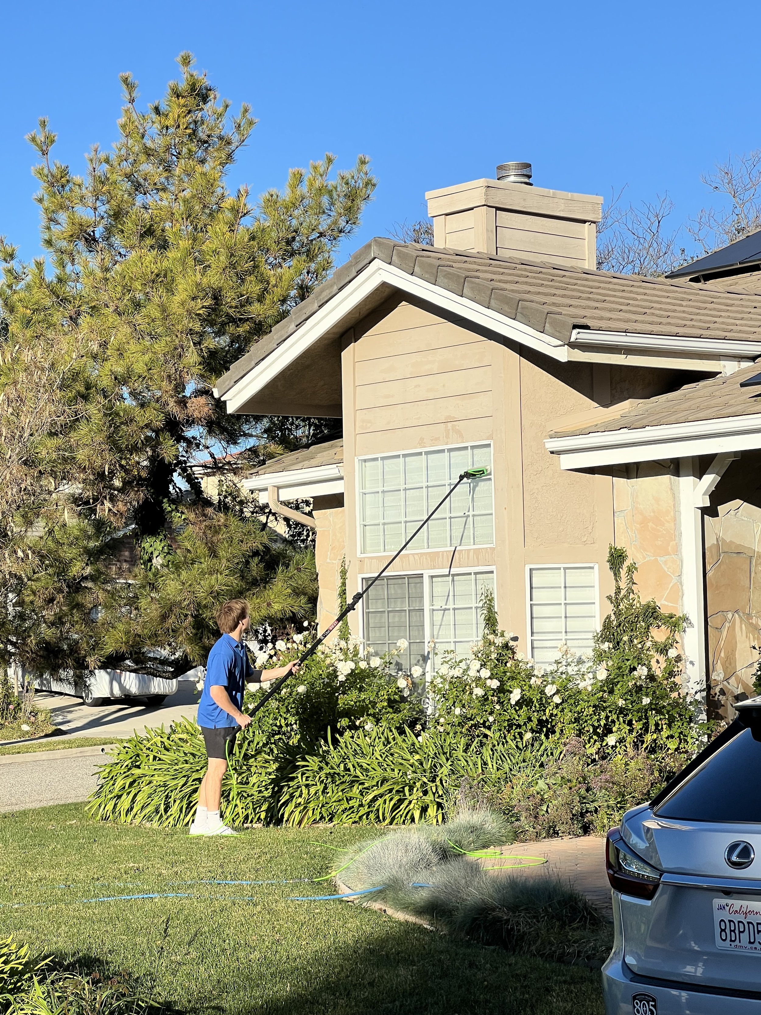 A person in a blue shirt and shorts is watering a garden with a hose in front of a house with a brick and beige wall, windows, a chimney, and greenery, including a large tree and bushes, under a clear blue sky.