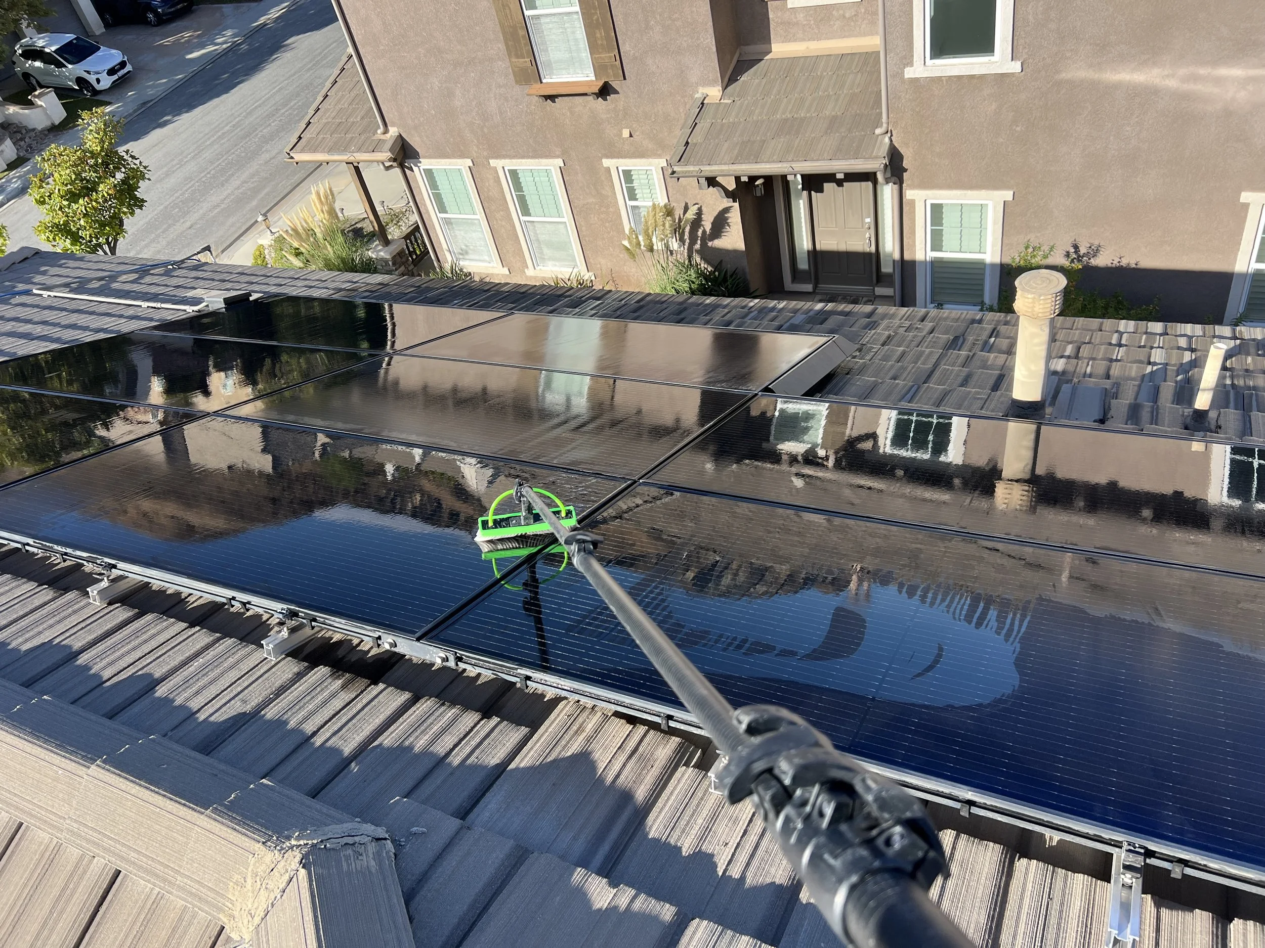 View from a rooftop showing solar panels, a house roof, and neighboring houses, with a person cleaning the solar panels using a long-handled tool.