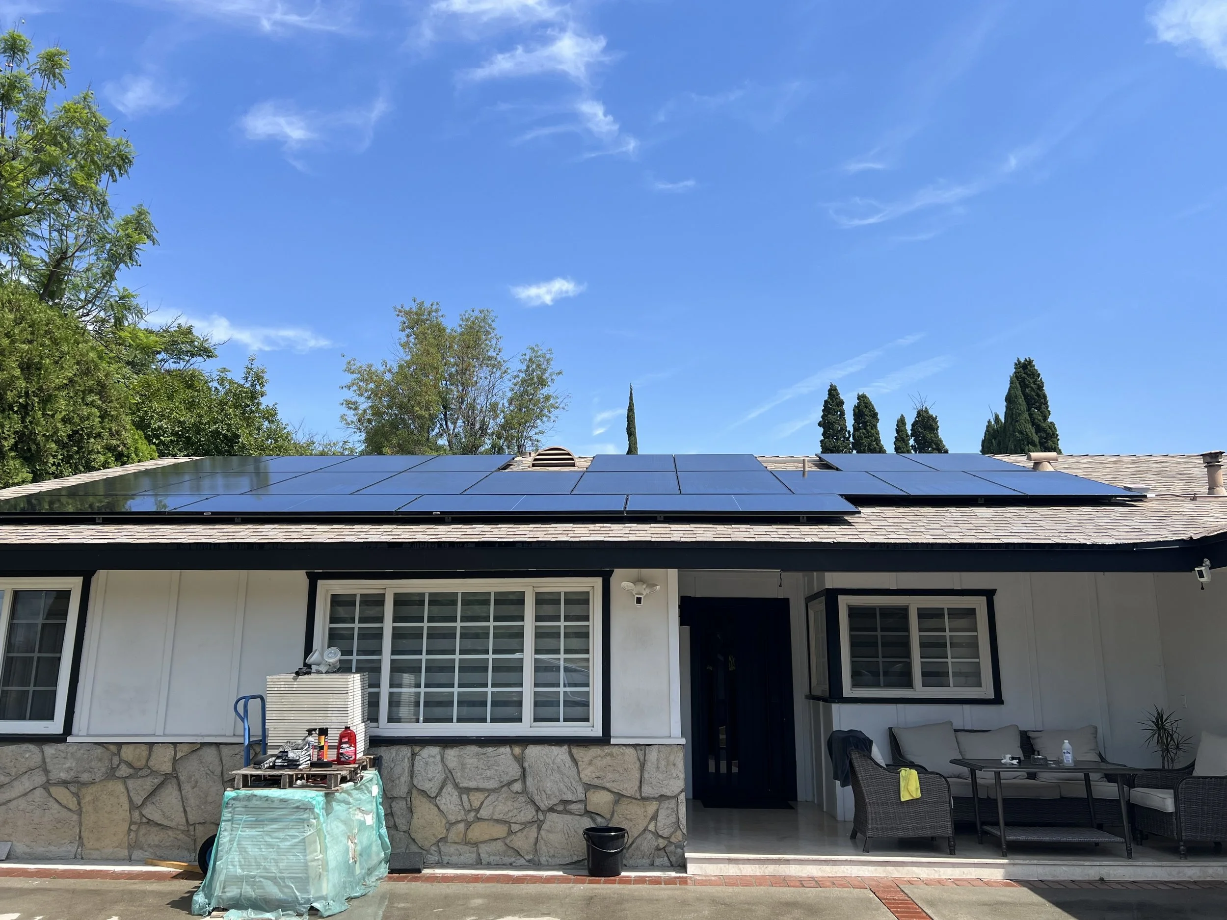 Backyard patio with roof-mounted solar panels, trees, and a blue sky with scattered clouds. Outdoor furniture, plants, and some equipment are visible.
