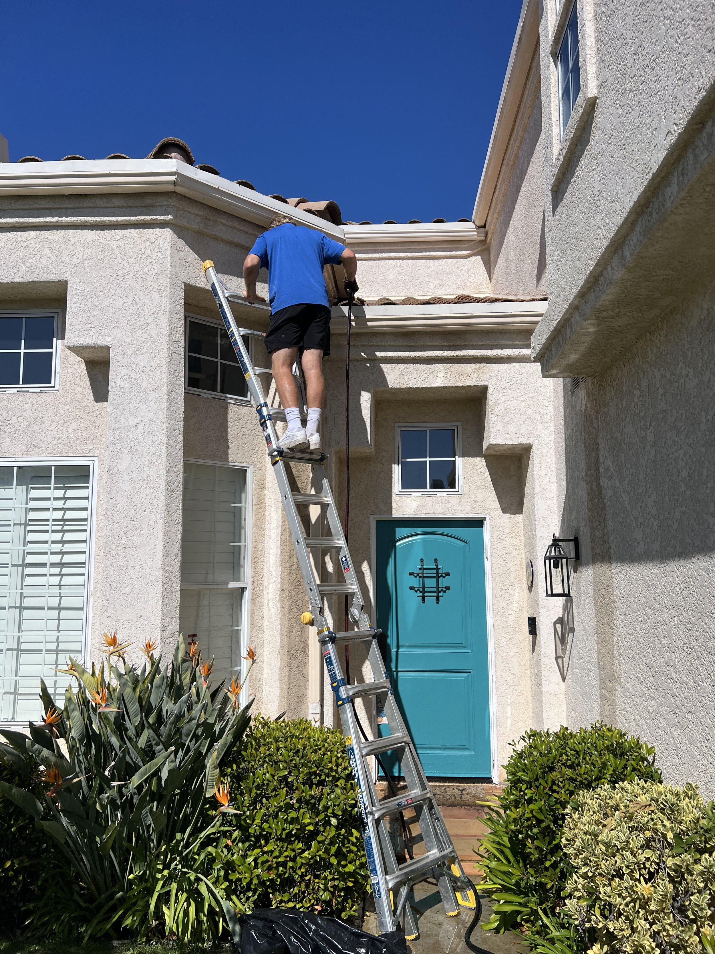 A person working on a ladder outside a house with a bright turquoise door, beige stucco walls, and windows, surrounded by green bushes and plants, under a clear blue sky.
