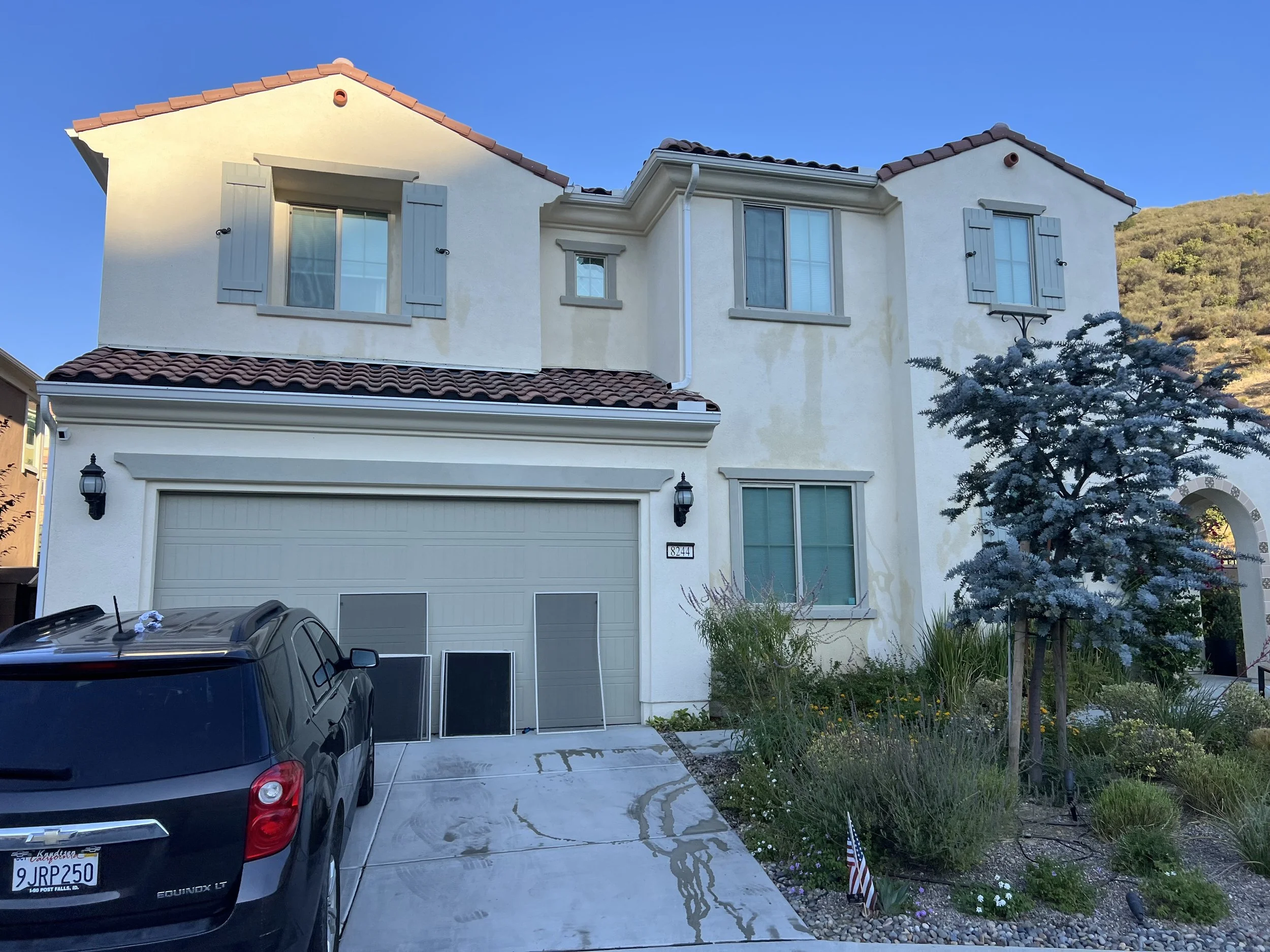 A multi-story house with a beige exterior, red-tiled roof, and several windows with shutters. A black car is parked in the driveway, and there is greenery with a garden and a tree with blue-green leaves in front of the house.